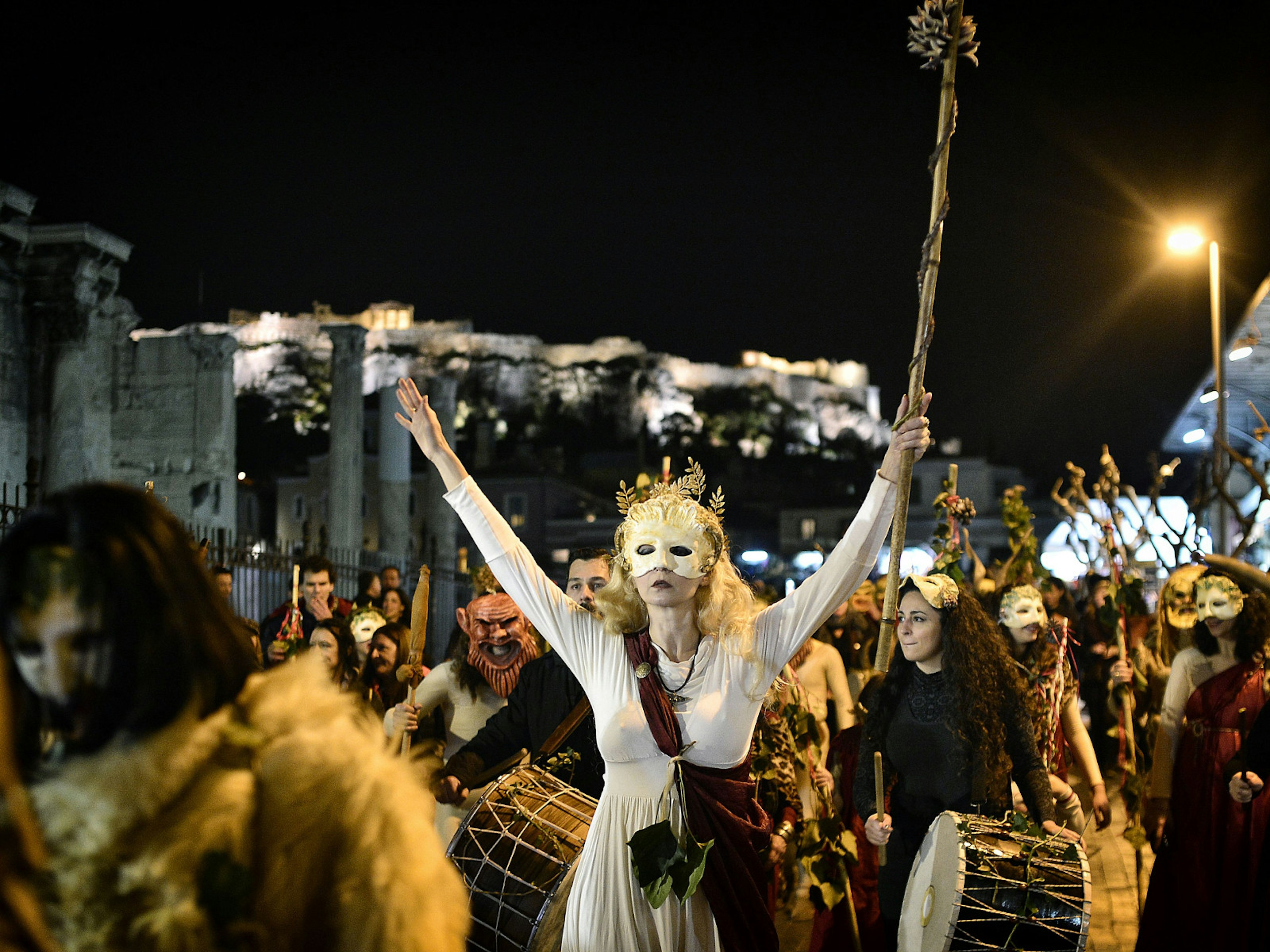 Heavily costumed people walk through the crowded streets. A masked woman holds her arms up in the air