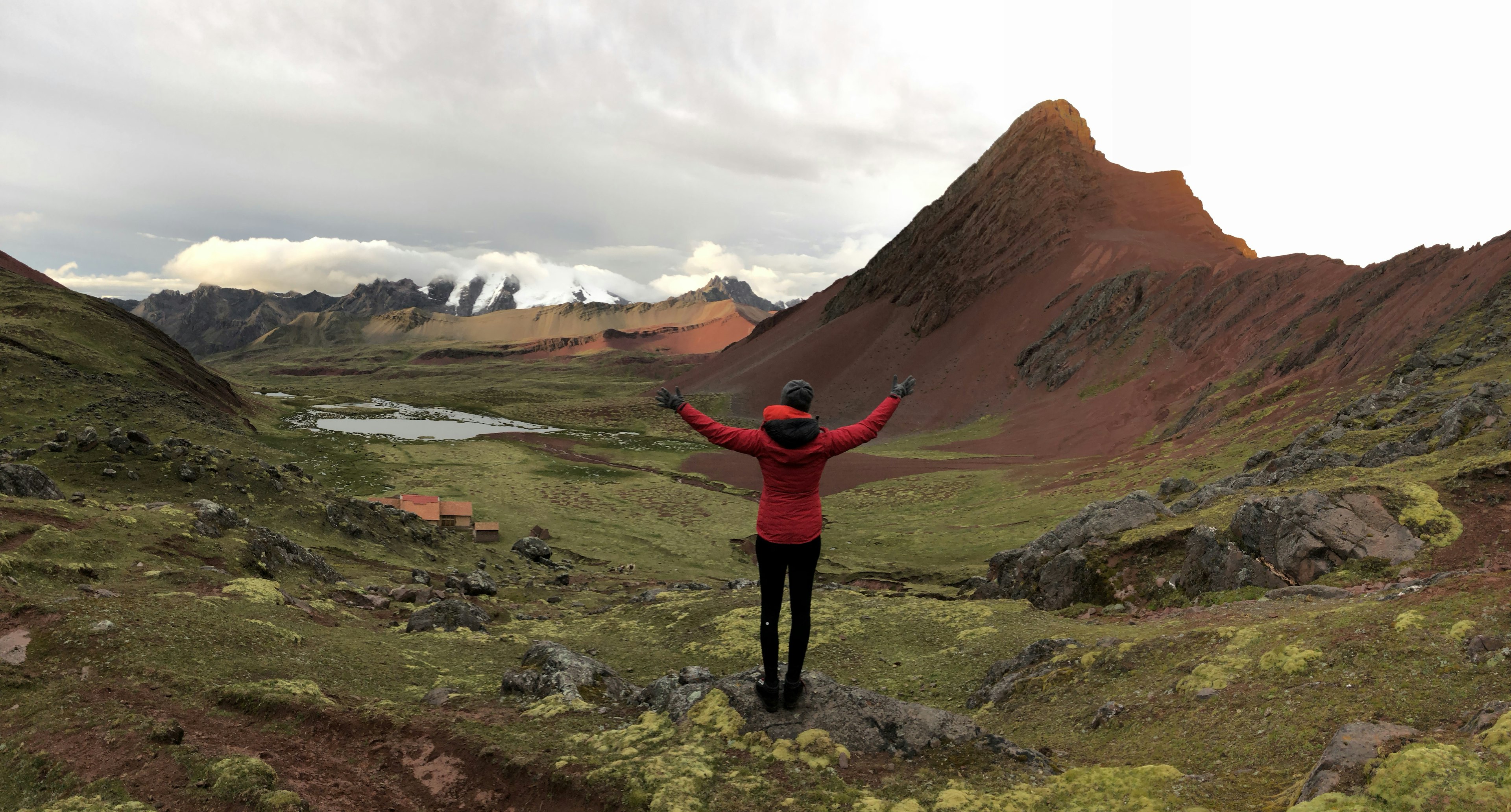 A woman in a red and black jacket extends her arms as she looks over a vast mountain landscape.