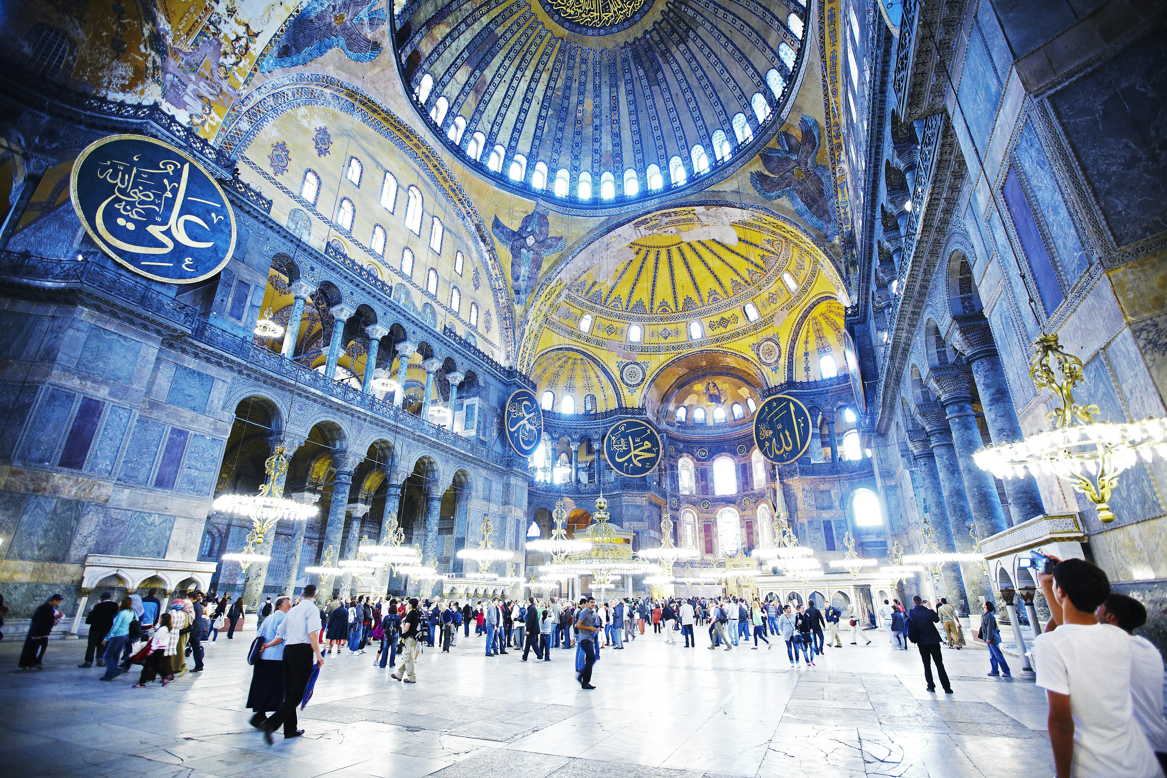 The light blue interior of Aya Sofya is highlighted with golden accents throughout the structure. There are large blue and gold signs attached to pillars around the building.