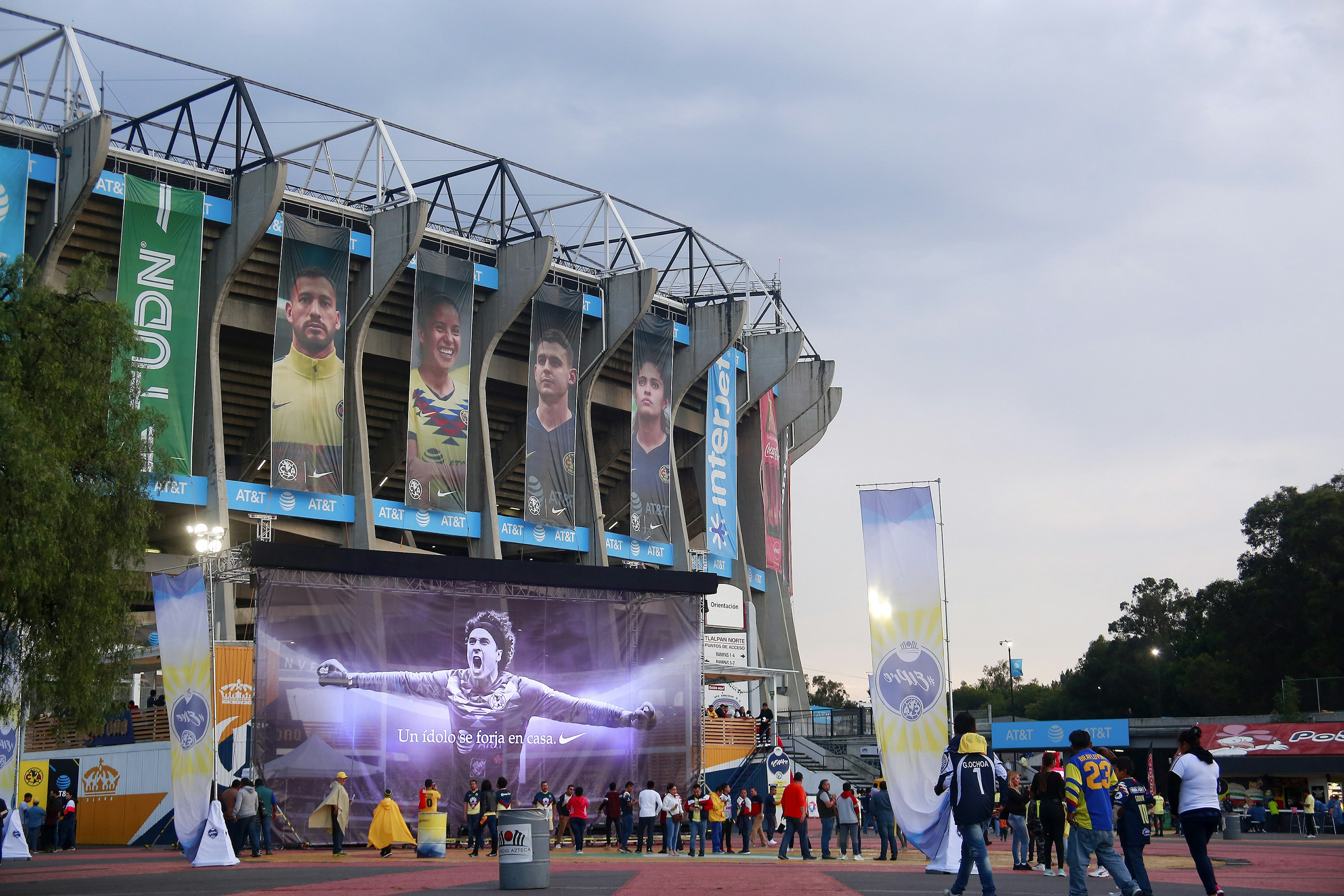 Large banners with the faces of soccer players hang on the side of Azteca Stadium. There are large groups of people heading to the stadium.