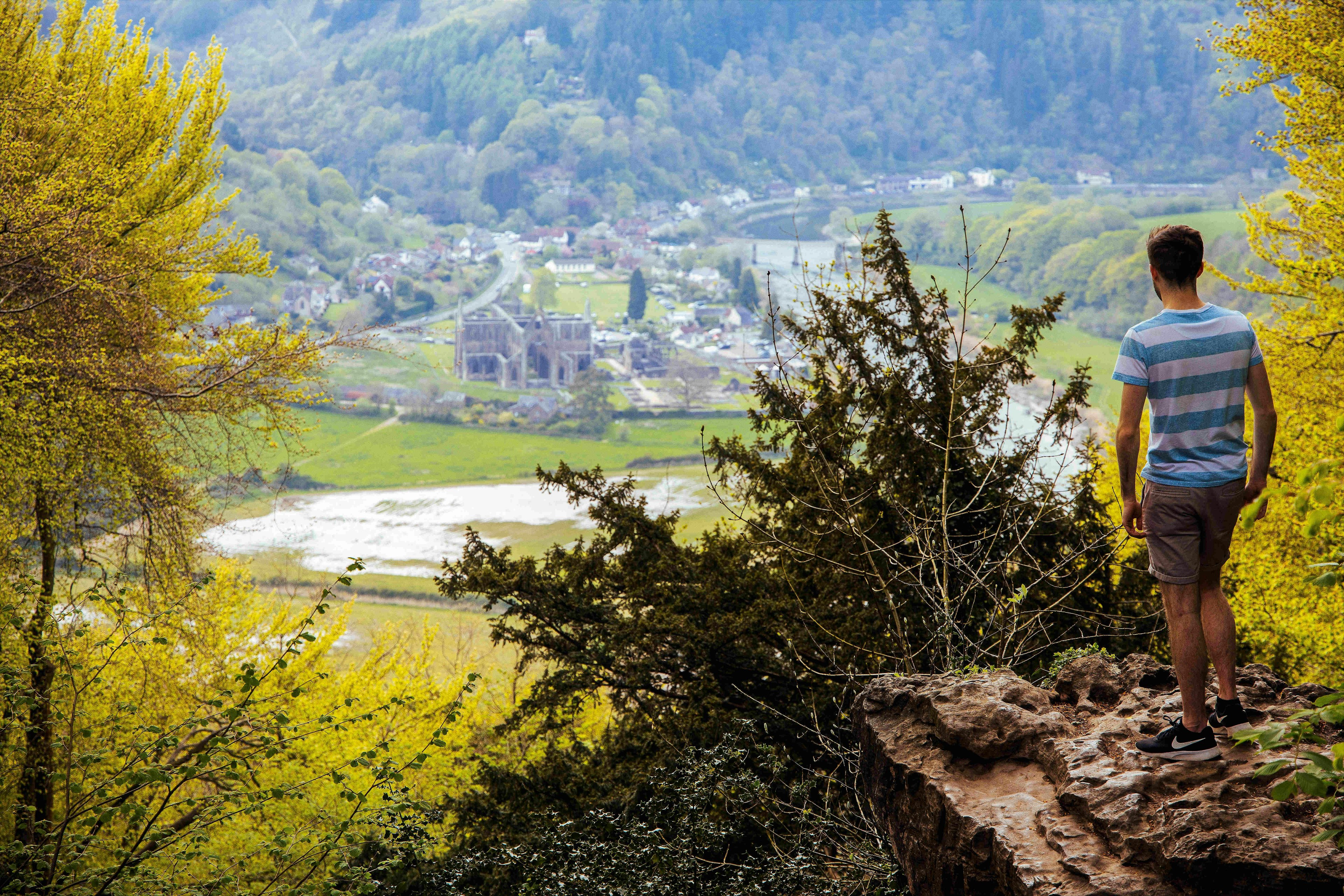 A man stands on a rocky ledge looking down on the ruins of Tintern Abbey