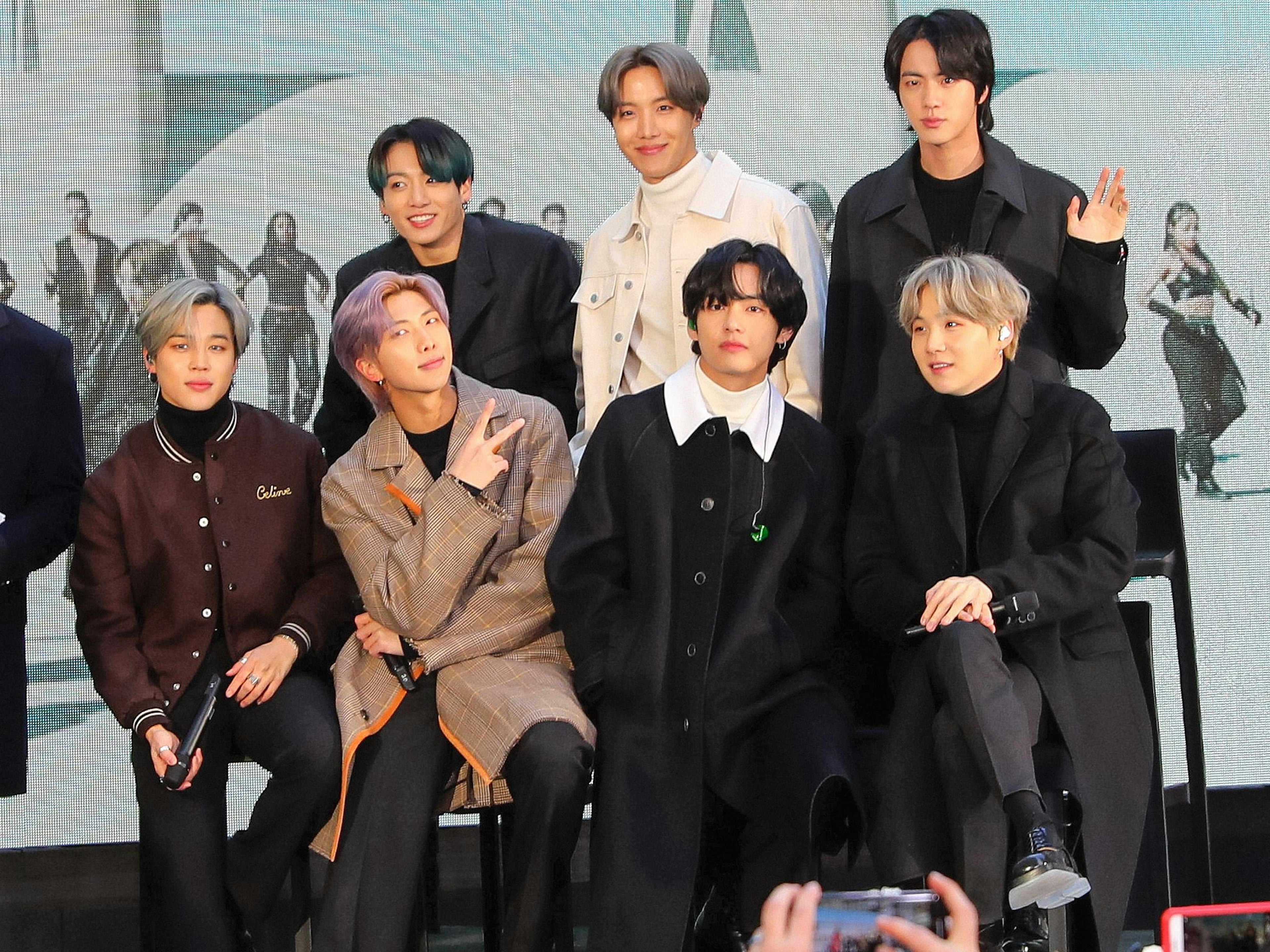 The seven members of K-Pop boy band BTS pose in front of a video screen.