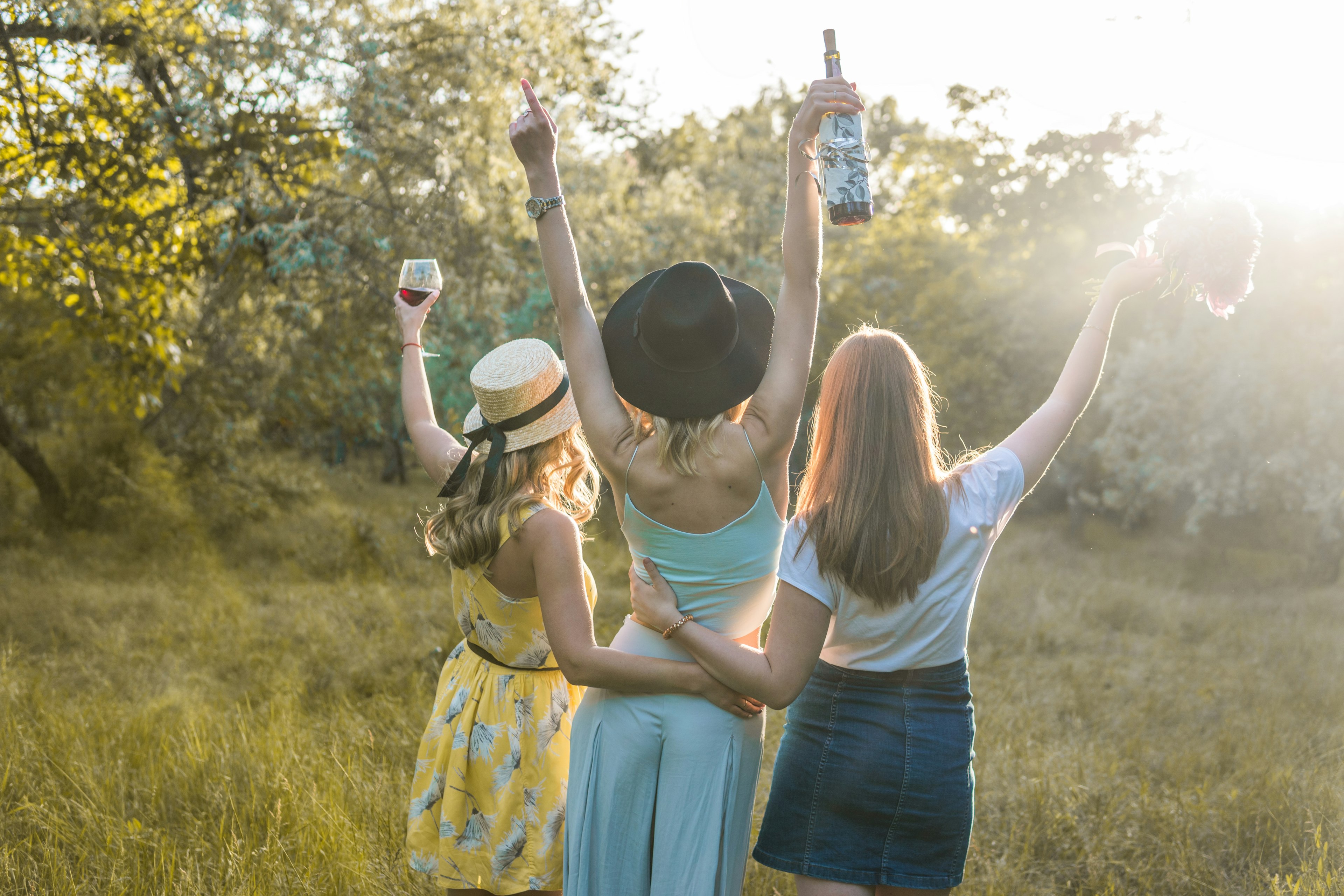 A trio of women with their backs facing the camera stand in the middle of a field, facing the sun. One woman is raising a wine glass, another is raising a wine bottle and another is holding a bouquet of flowers; bachelorette party