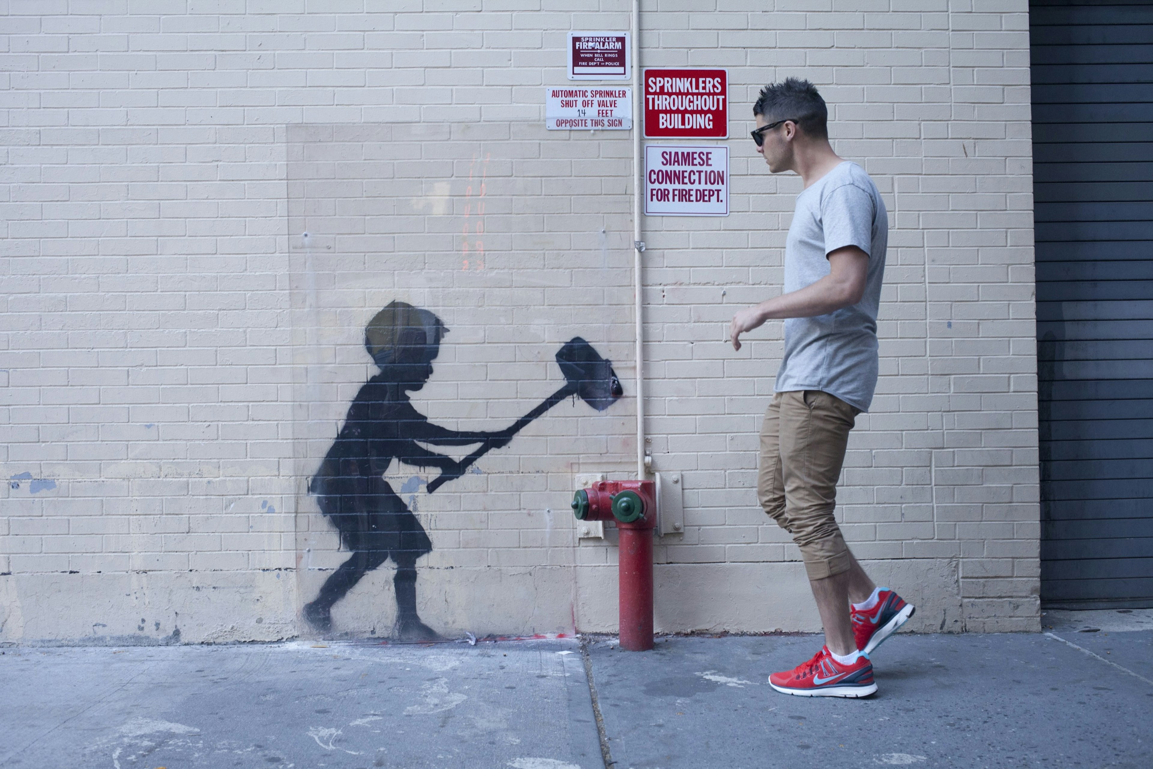 A man walks past a stenciled artwork on a building in New York City, of a boy wielding a hammer