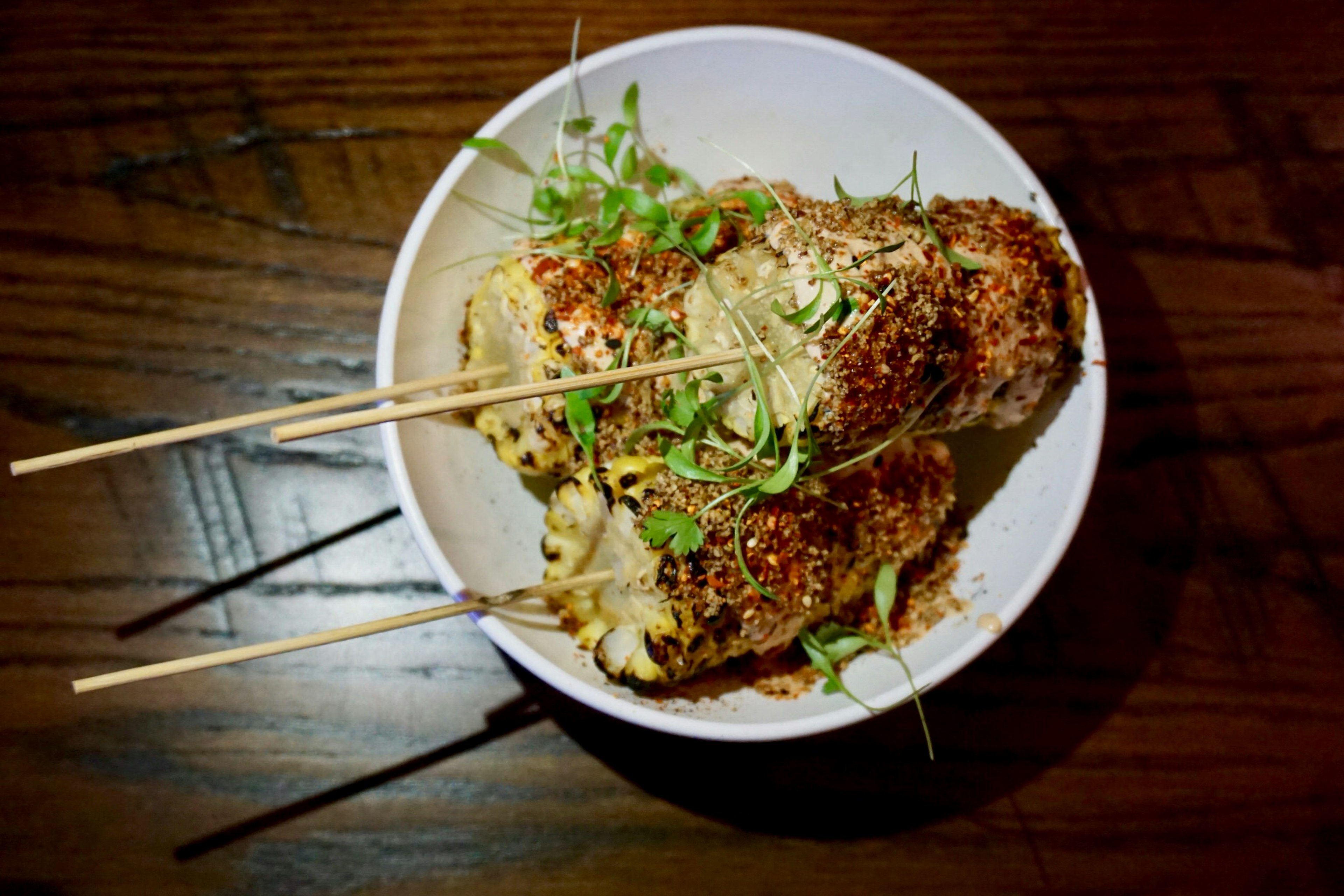 A white bowl on a dark wooden table containing three liberally seasoned corn-cobs, each with a wooden skewer sticking out of them. The dish is decorated with fresh herbs.