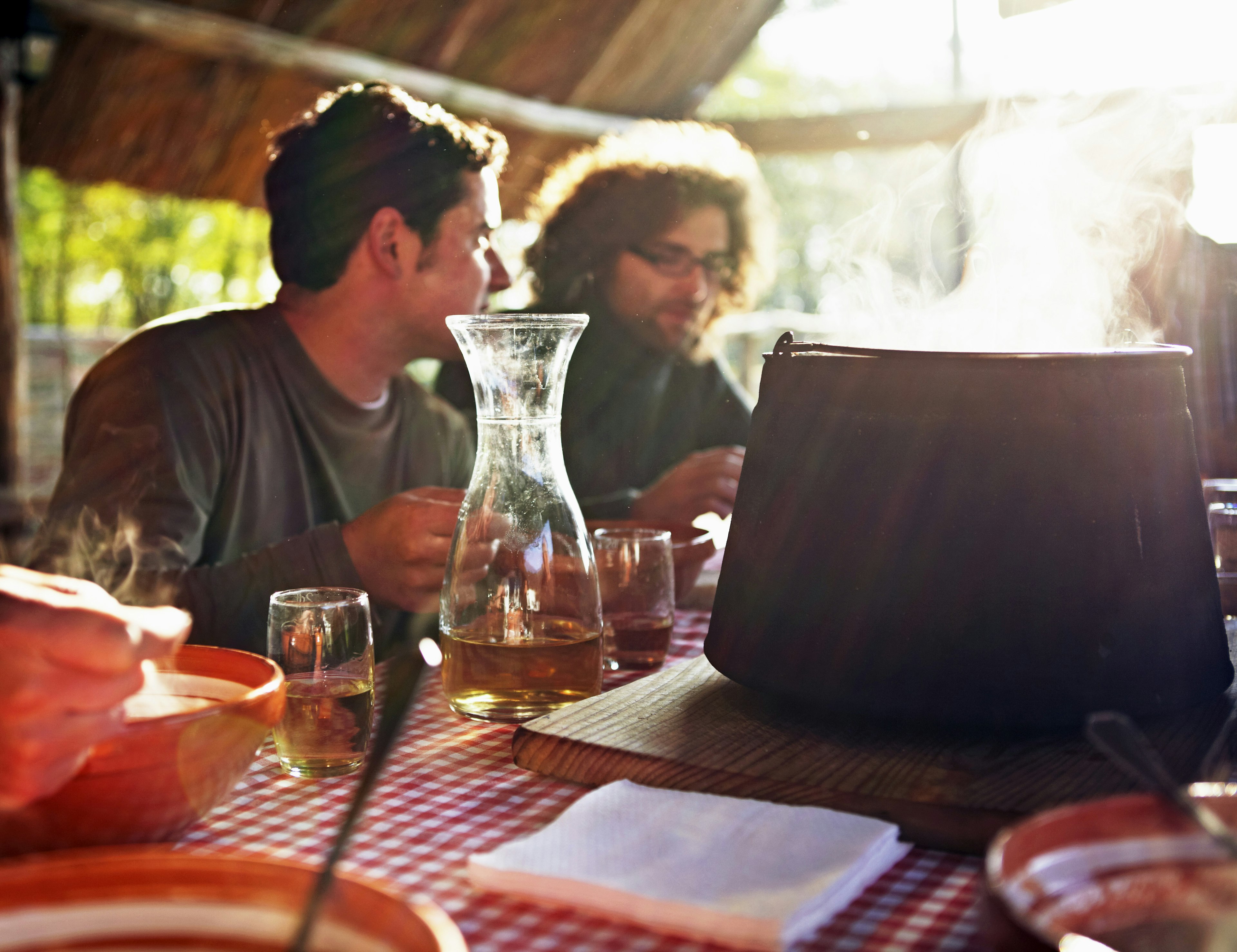 A group of friends sit around a red-checkered table with a steaming pot in the center of it