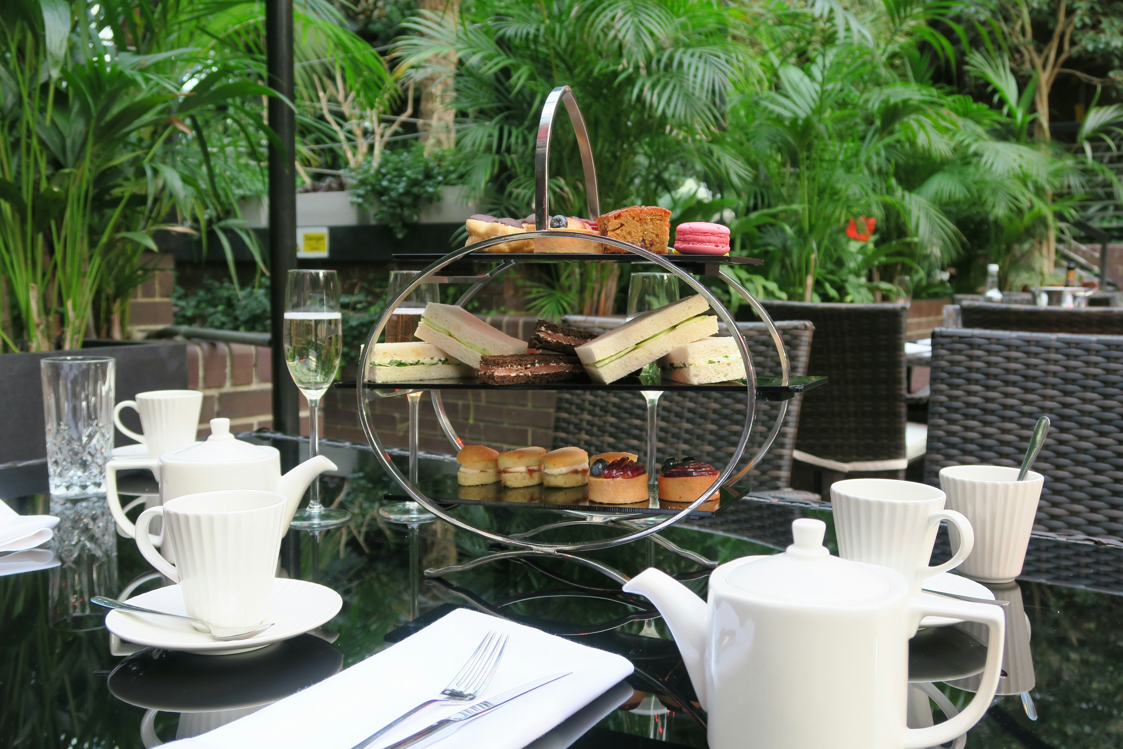 White cups and teapots on a table with a platter of cakes and sandwiches and ferns and palms in the background.