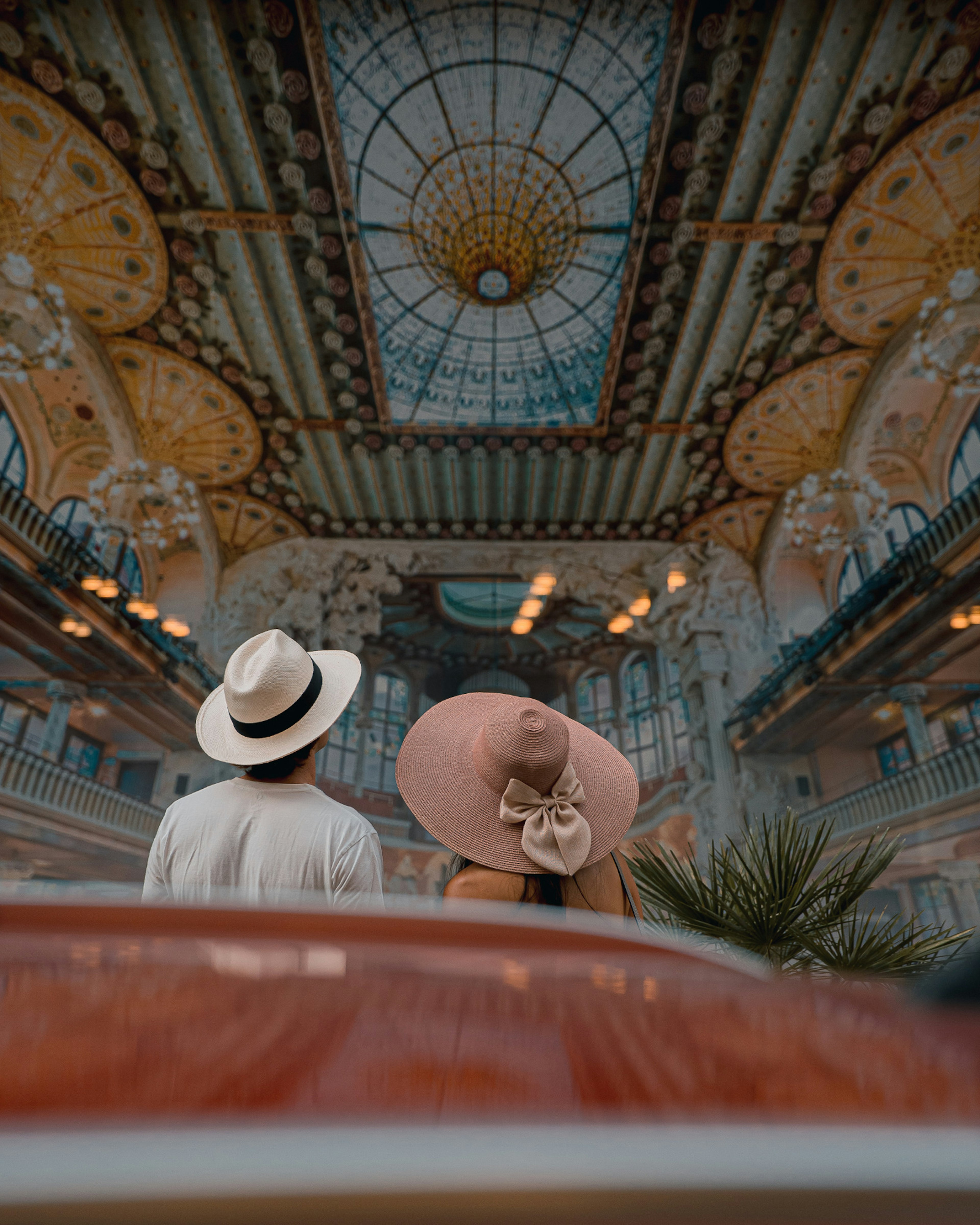 A couple in the Palau de la Música Catalana in Barcelona, Spain; it is a richly colourful auditorium, and they gaze upwards at a ceiling of blue-and-gold stained glass and a shimmering skylight that looks like a giant, crystalline, downward-thrusting nipple.