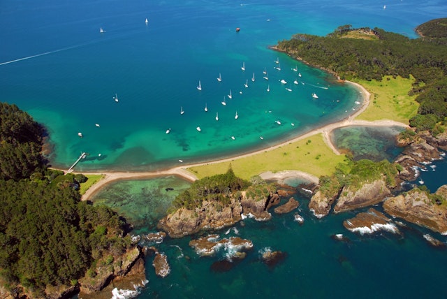 Aerial shot of boats off Roberton Island, Bay of Islands, New Zealand; the boats are all at achor in a horseshoe shaped bay with torquoise waters near the beach it its heart; the ends of the bay are both forested; the opposite side to the bay across a narrow isthmus is a shore dominated by tall rocky outcrops topped by trees