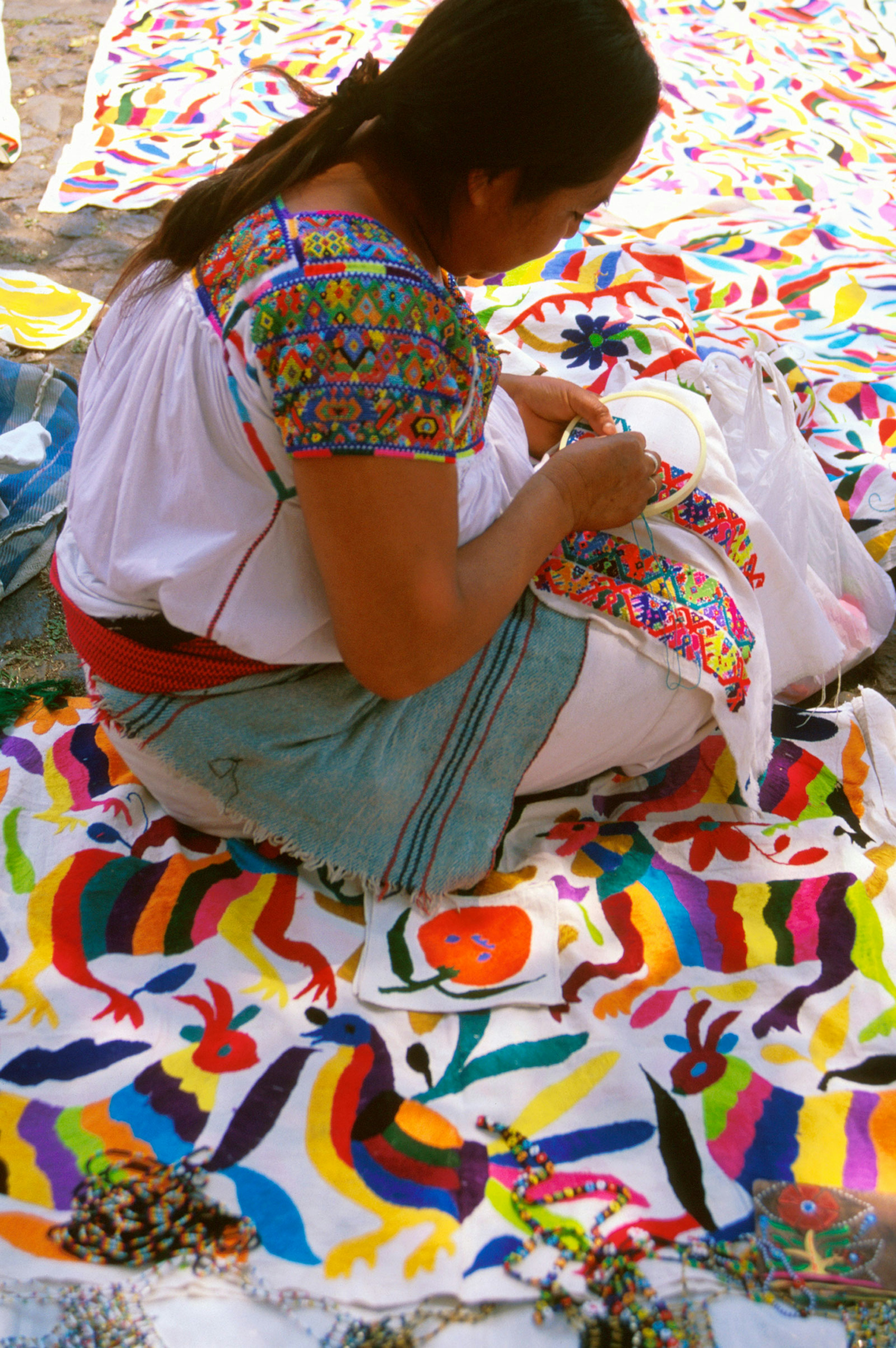 A woman embroiders colorful designs on a shirt while sitting on a colorful blanket