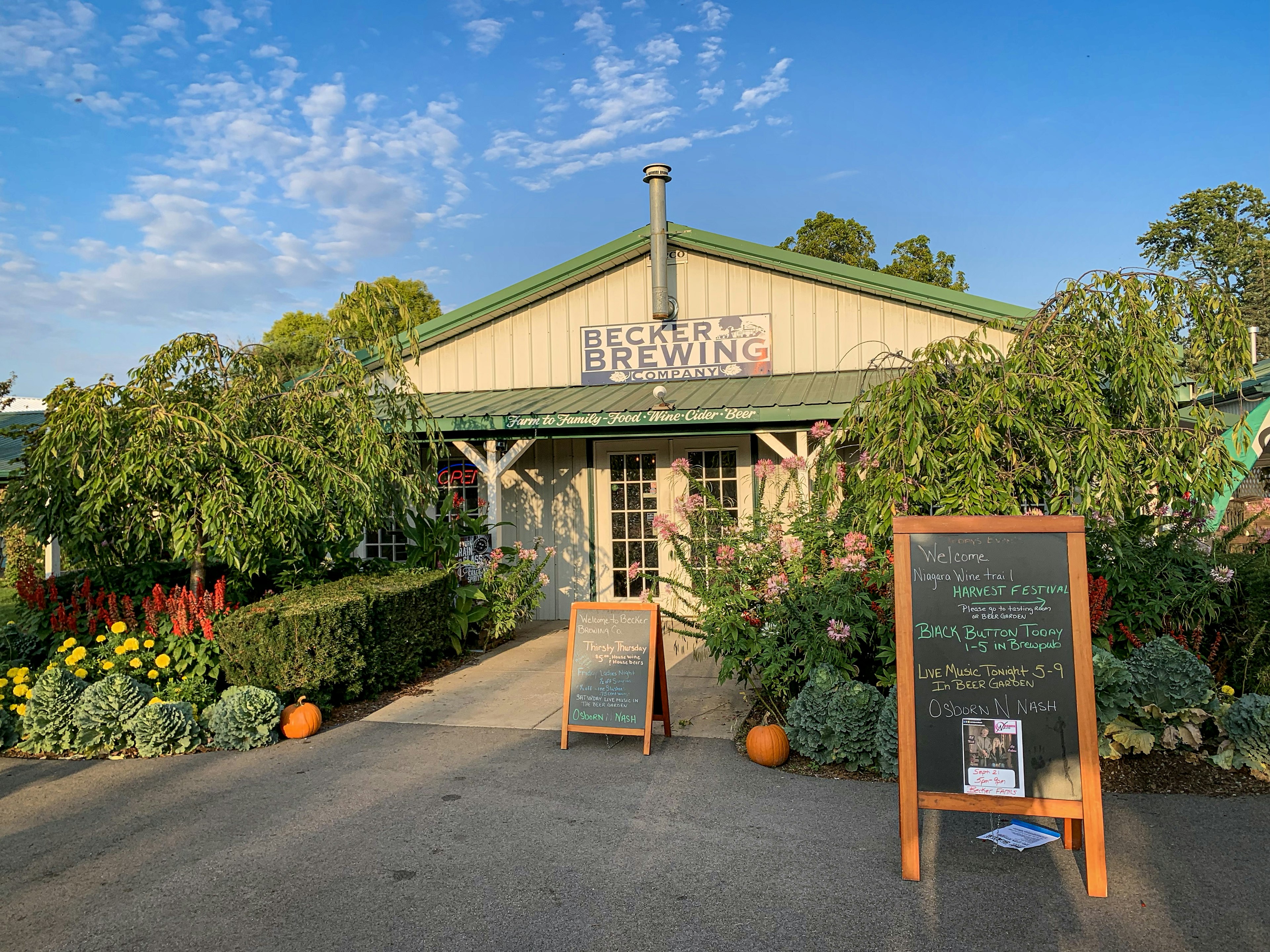 Exterior of Becker Farm features tall green plants and a pair of wooden sign. There are a pair of pumpkins on either side of the entranceway.