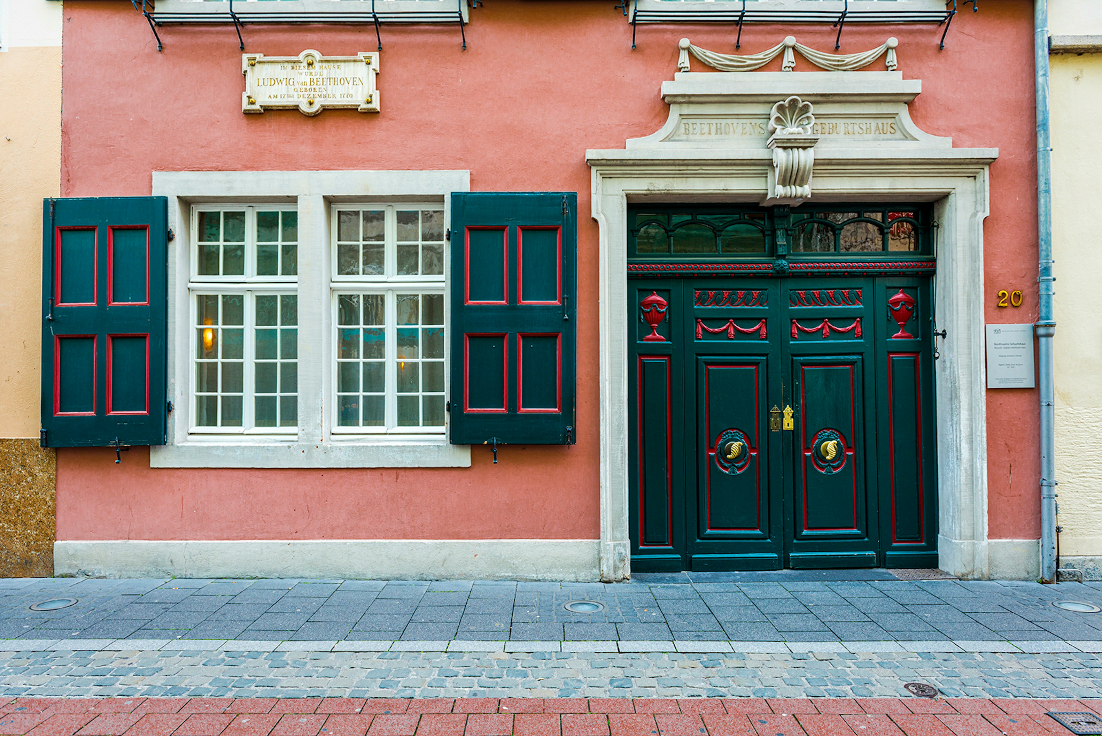 A salmon-colored cement house with an ornate door painted black with red trim and matching shutters on the window to the left of the door