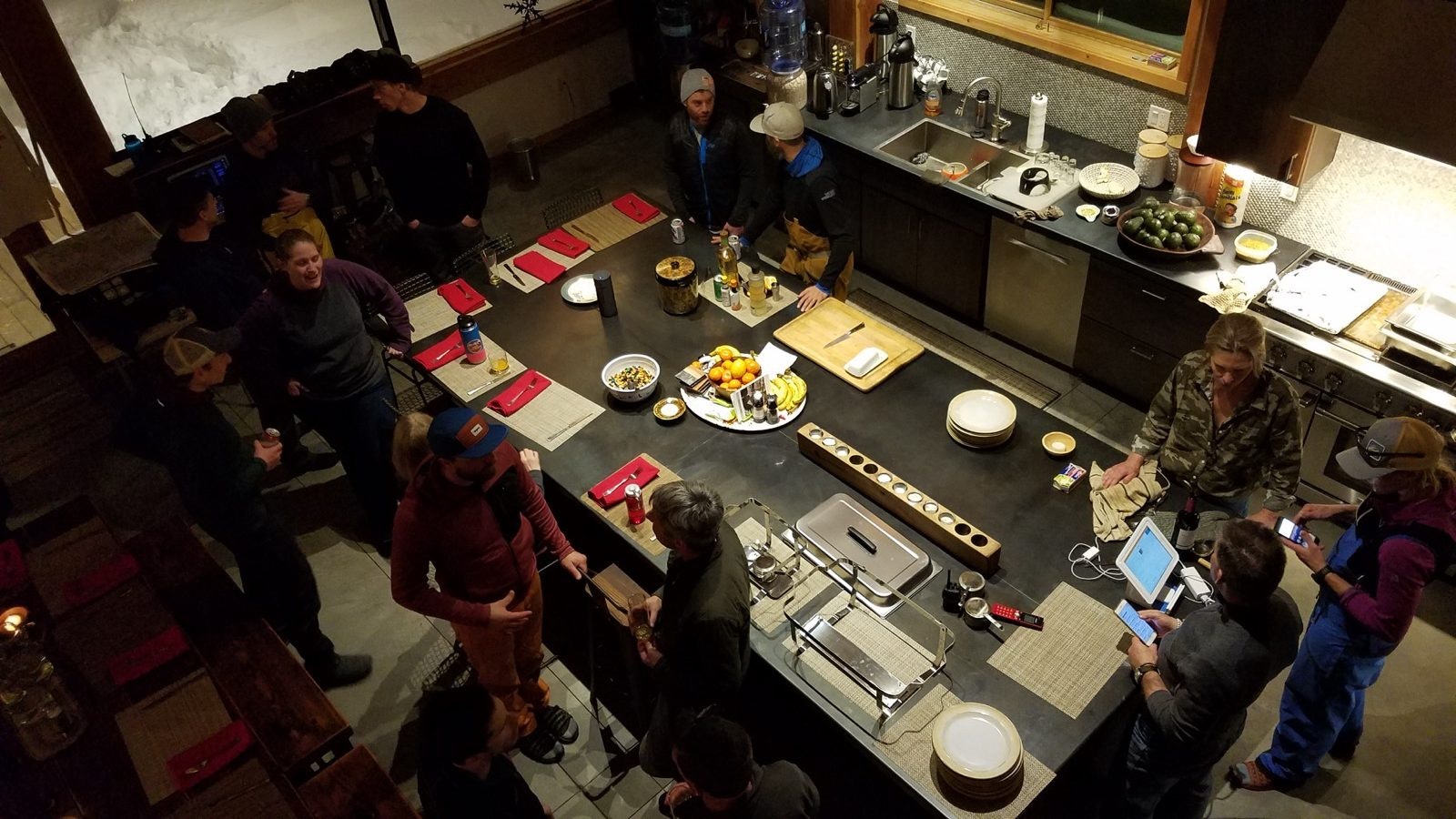 Looking down from above on a group of young people clustered around a large cooking and eating station in a lodge, where they are socializing