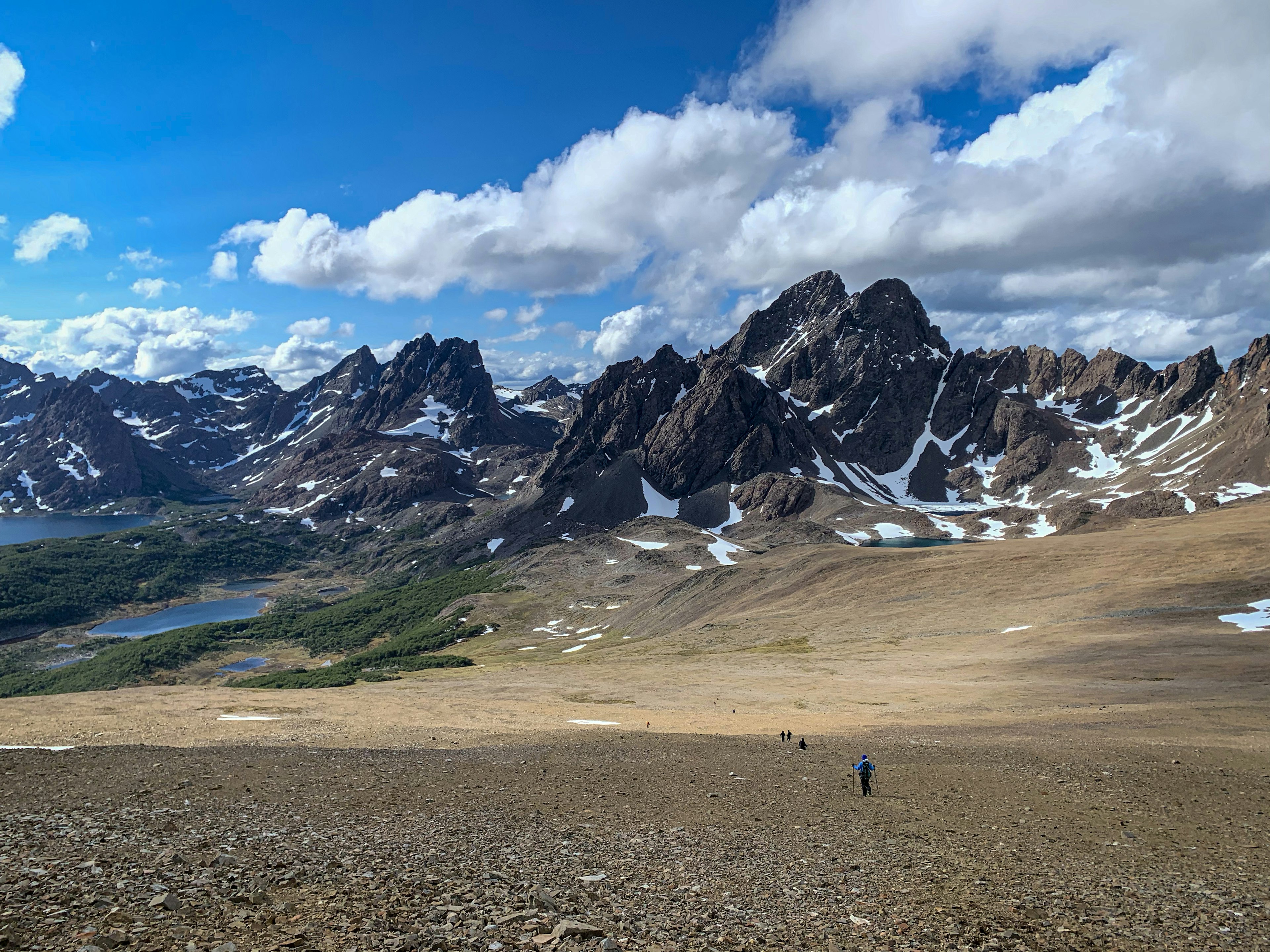 Hikers trek across loose ground, almost disappearing against the backdrop of the enormous, snow-capped, jagged mountains of the Dientes de Navarino.