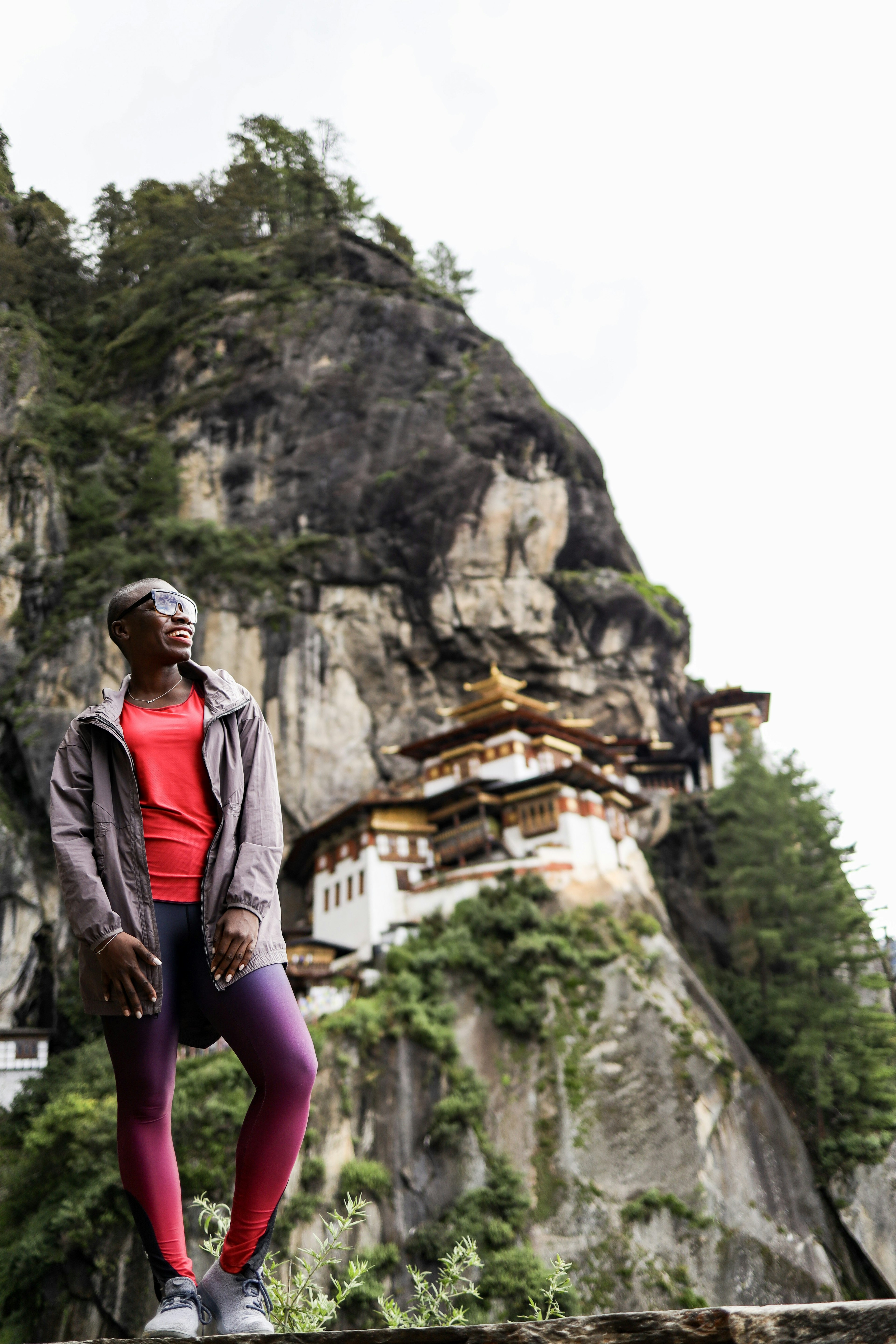 Jessica smiling looking away from the camera standing in front of a large house embedded in the side of a mountain in Bhutan