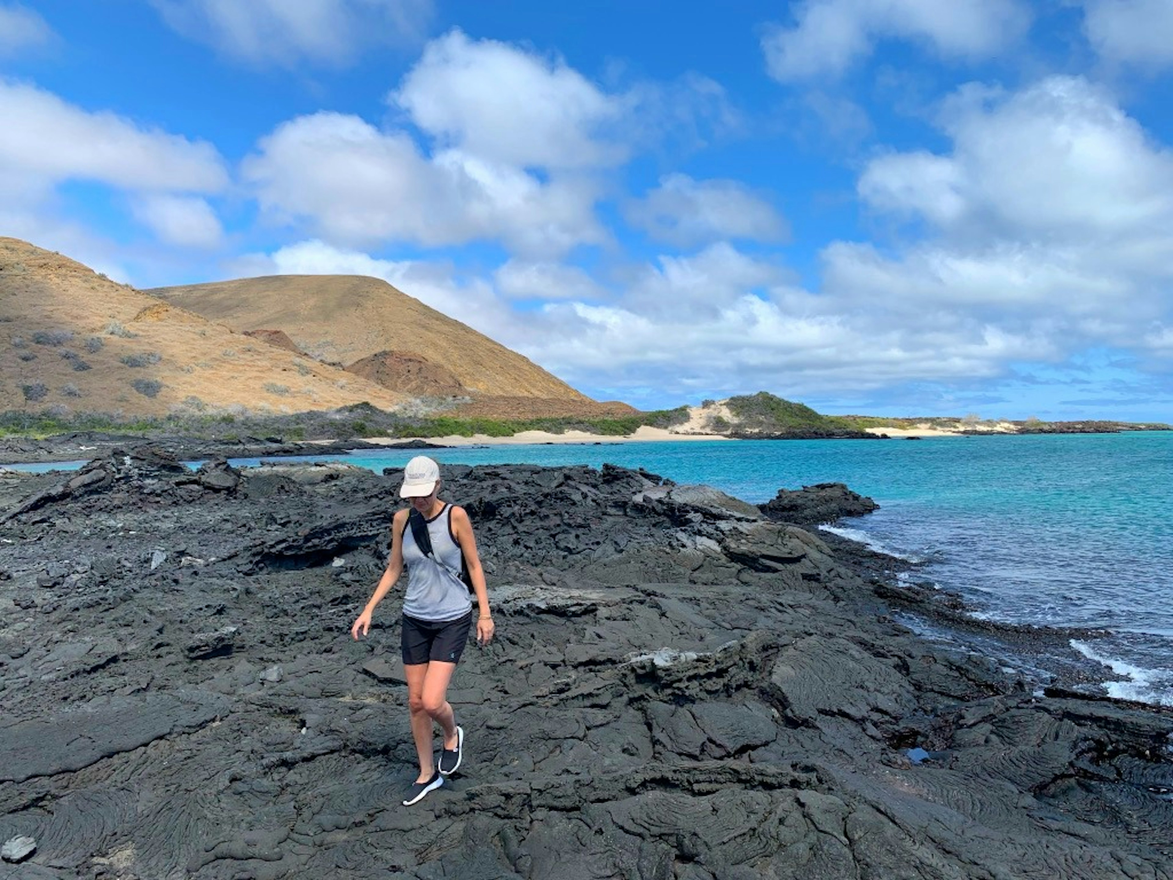 A woman wearing shorts and a tank top walks on black lava near the ocean in Galapagos