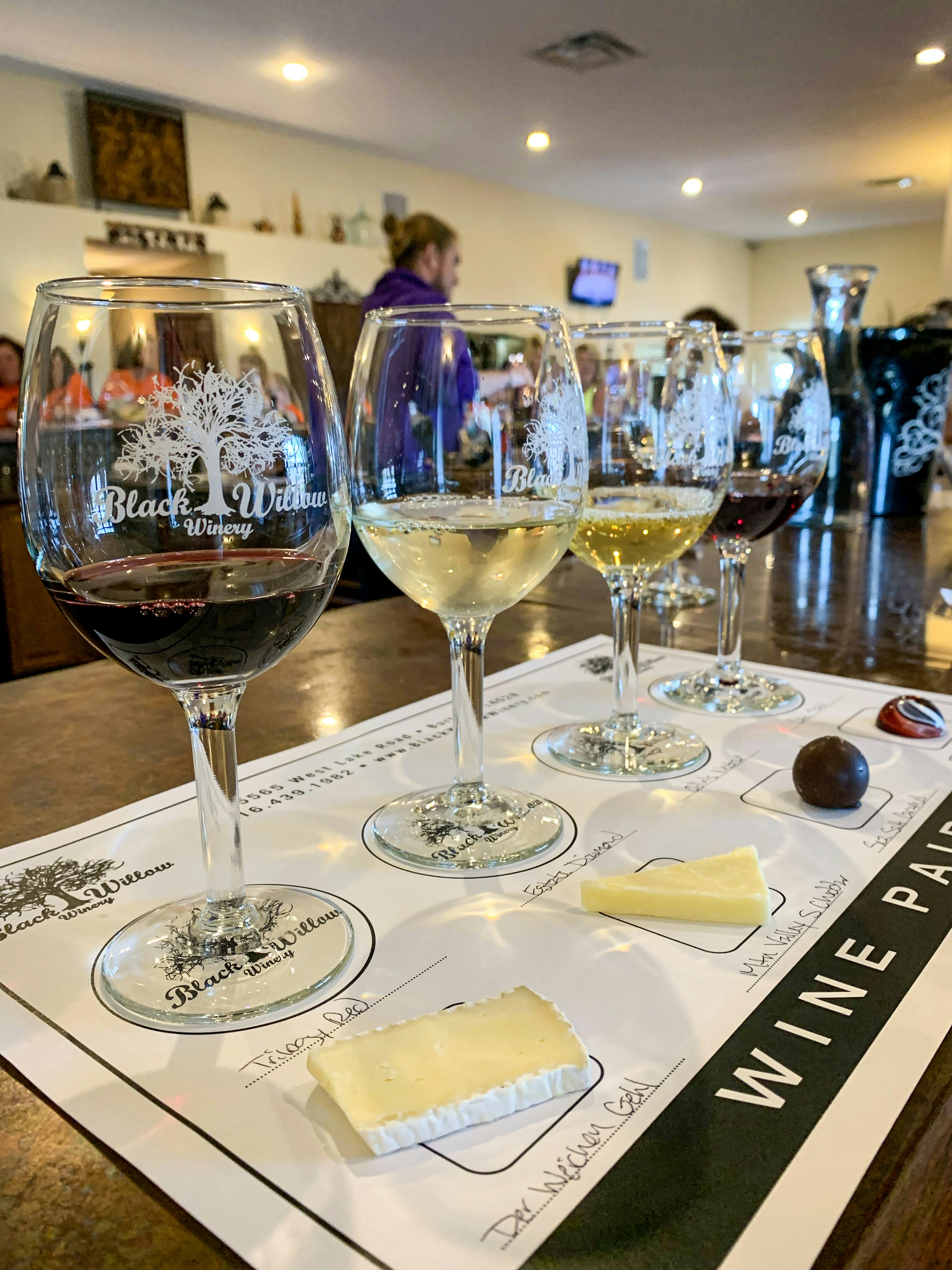 Four wine glasses, each filled with a different type of wine sit in front of an assortment of pairing items like cheese and chocolate in the Black Willow Winery