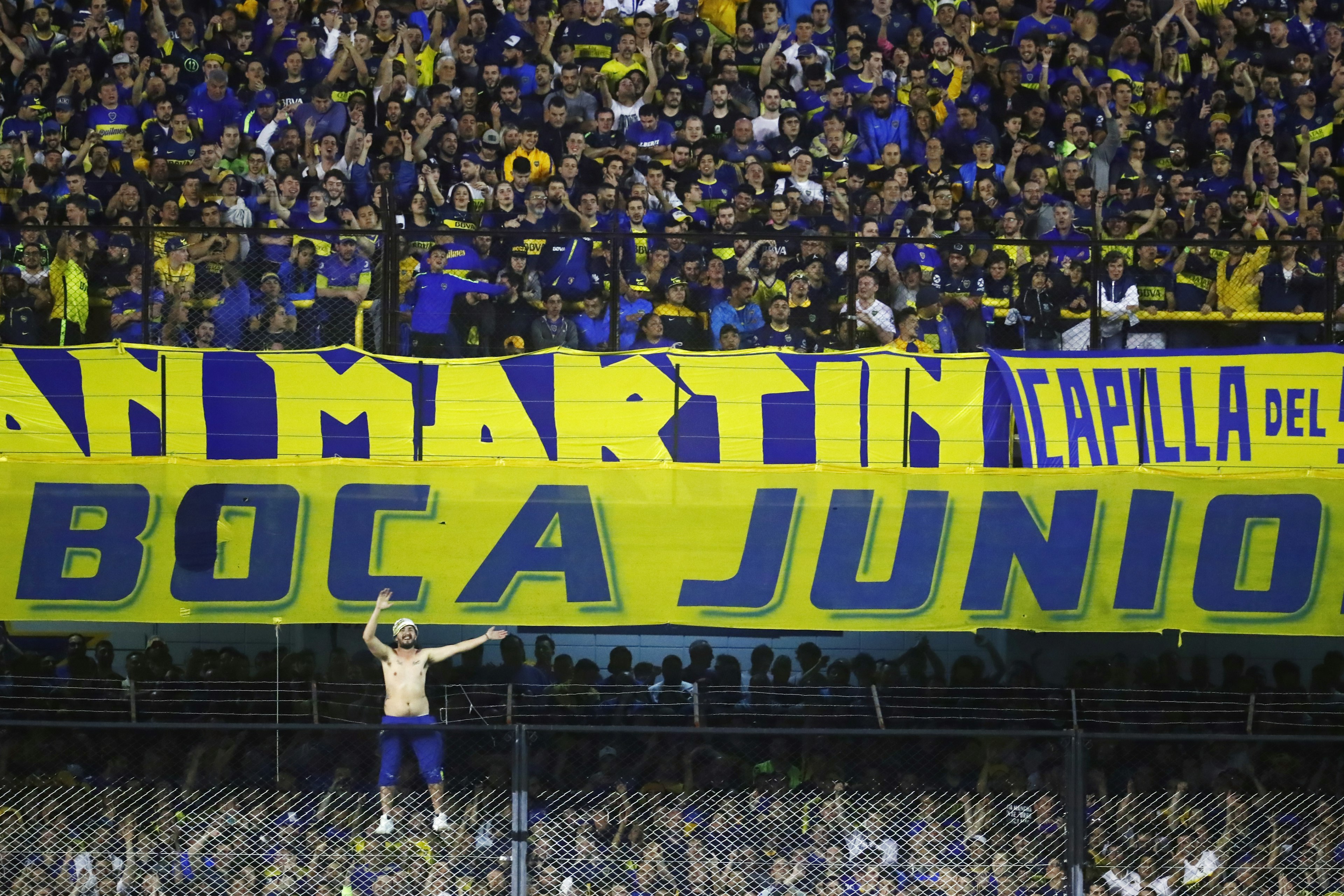 A shirtless man scales to the top of a fence topped with barbed wire at a soccer stadium in a sea of fans wearing blue and yellow in support of Boca Juniors