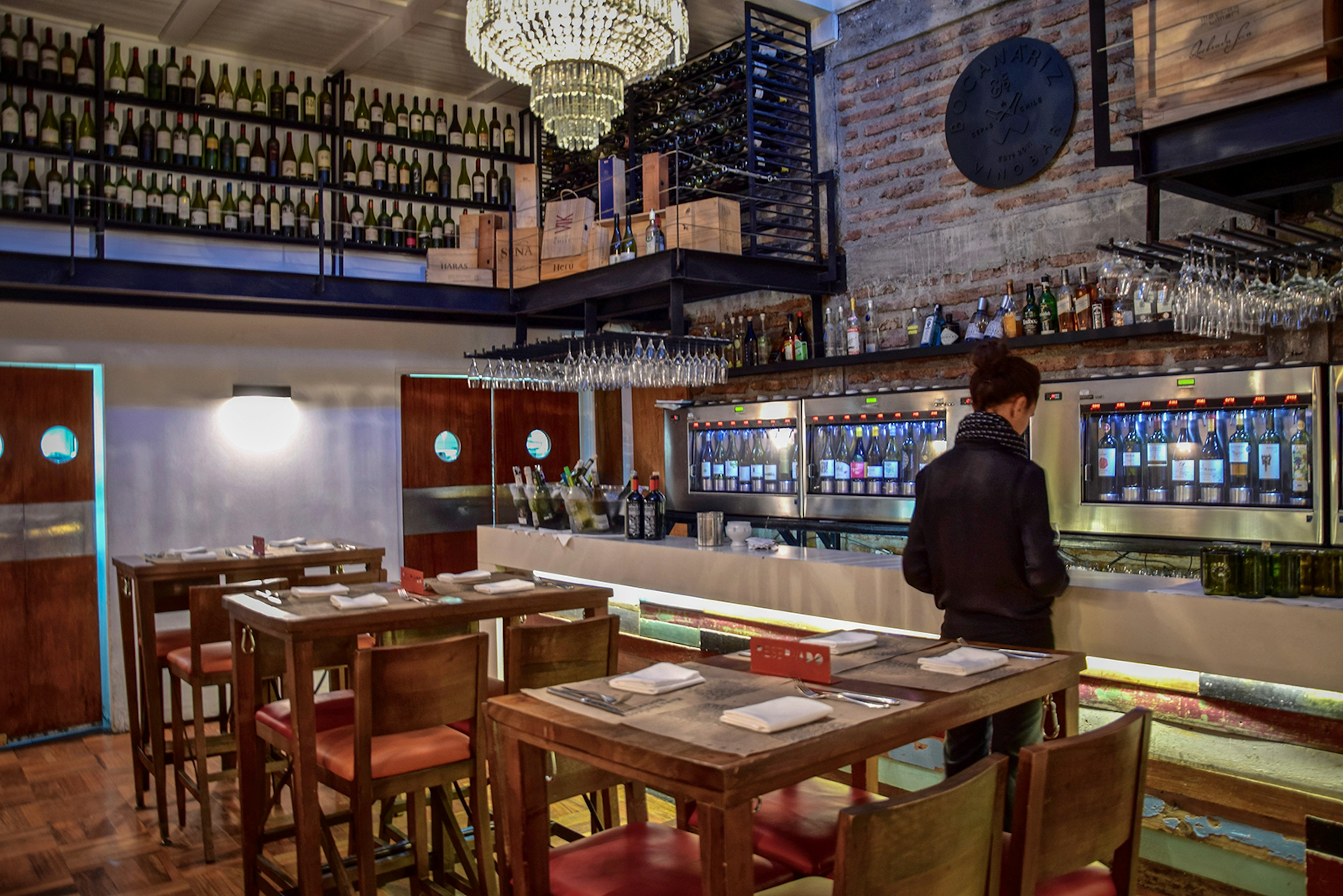 A woman stands at a bar counter with a wine cooler behind it; bottles of wine also line the bar wall