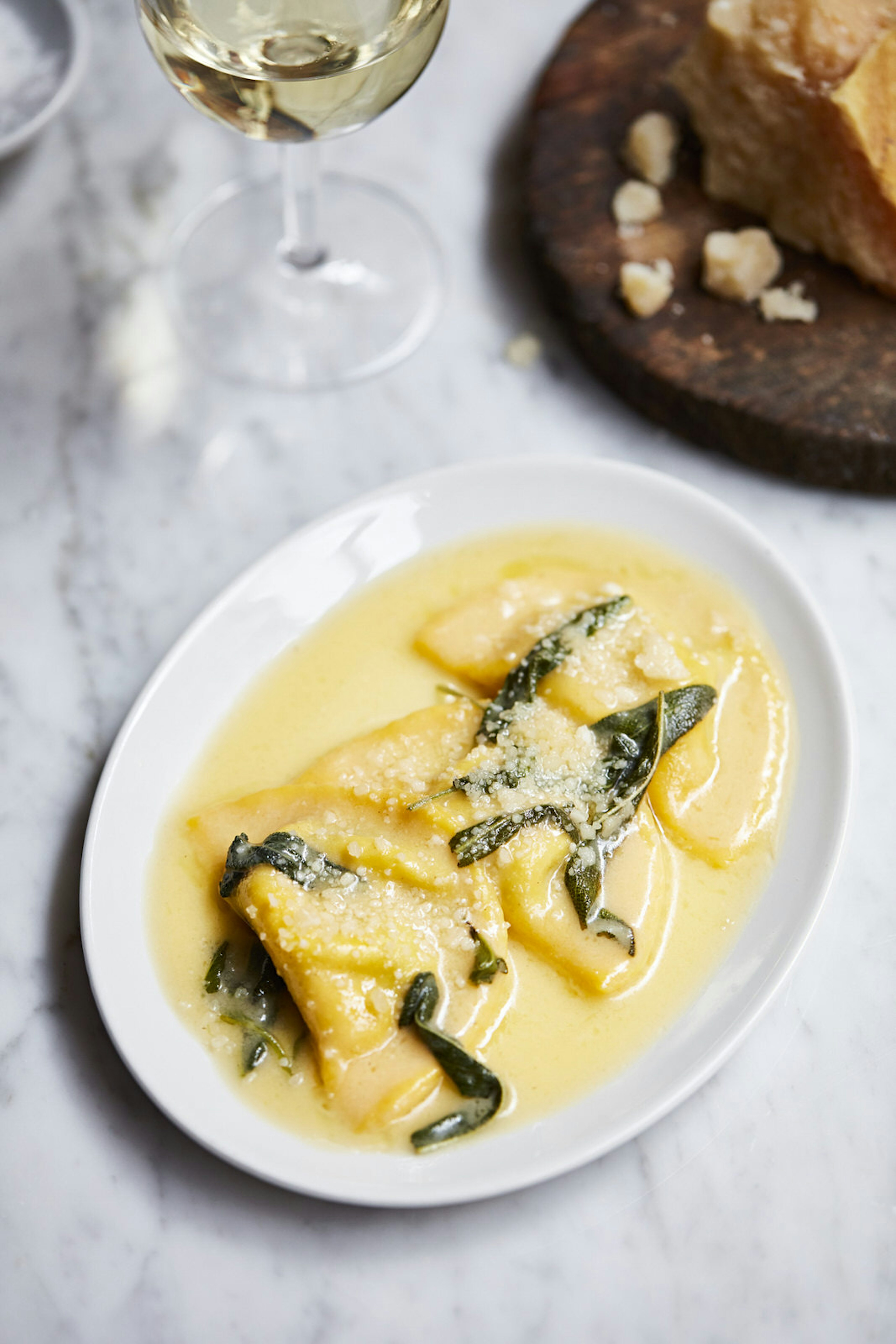 Borage and ricotta pansotti with butter and sage sit in an oval plate on a marble-topped table alongside a glass of white wine