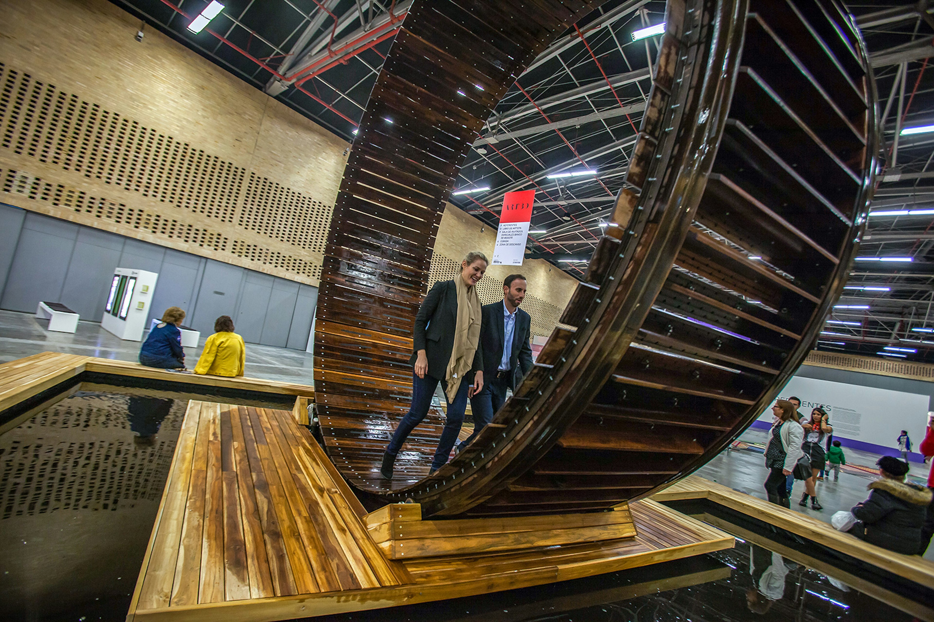 A man and a woman walk on what looks like a giant hamster wheel at an art exposition