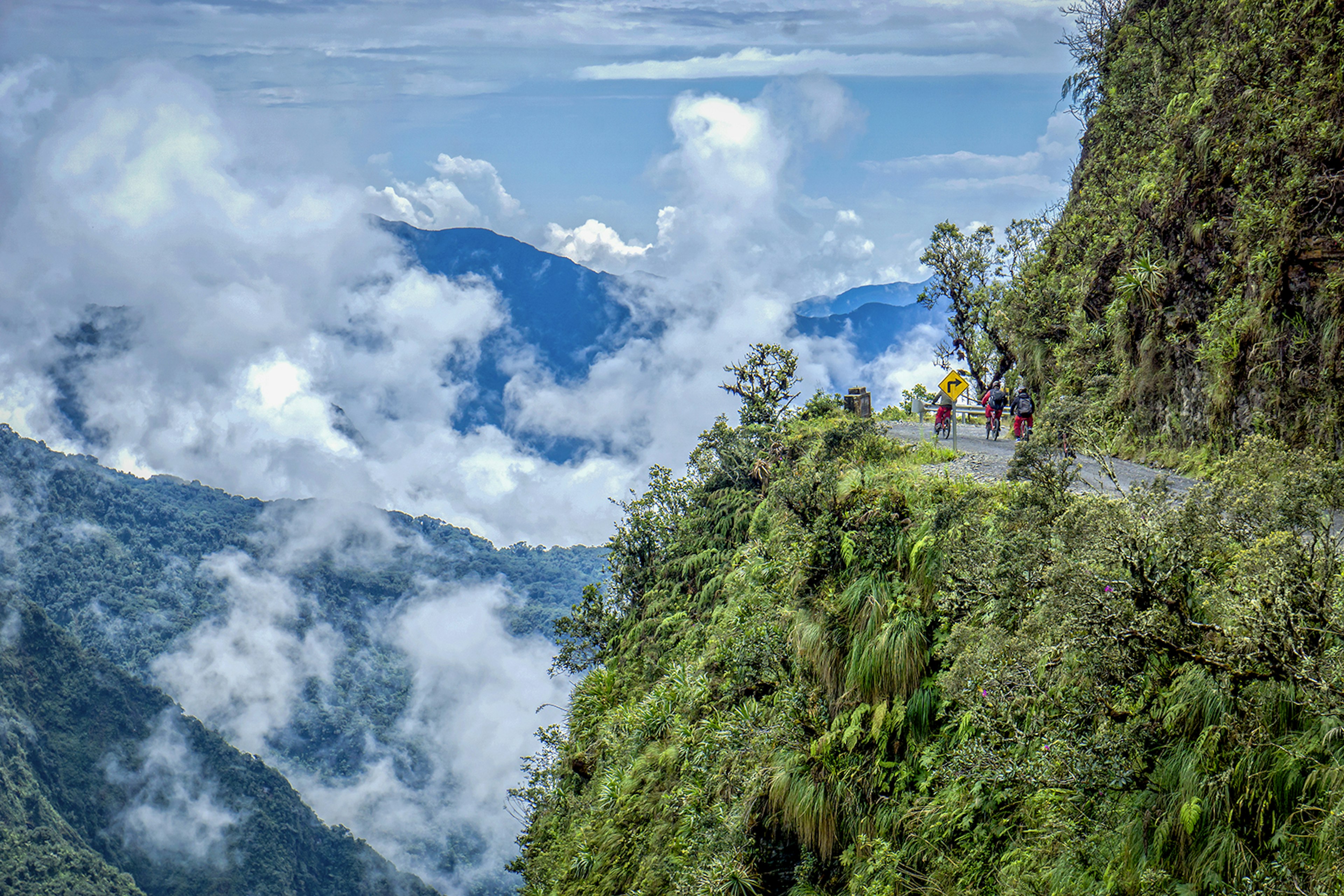 Cyclists on a small road on the side of a foliage-covered mountain, with a sheer drop-off to the left © Filrom / Getty Images