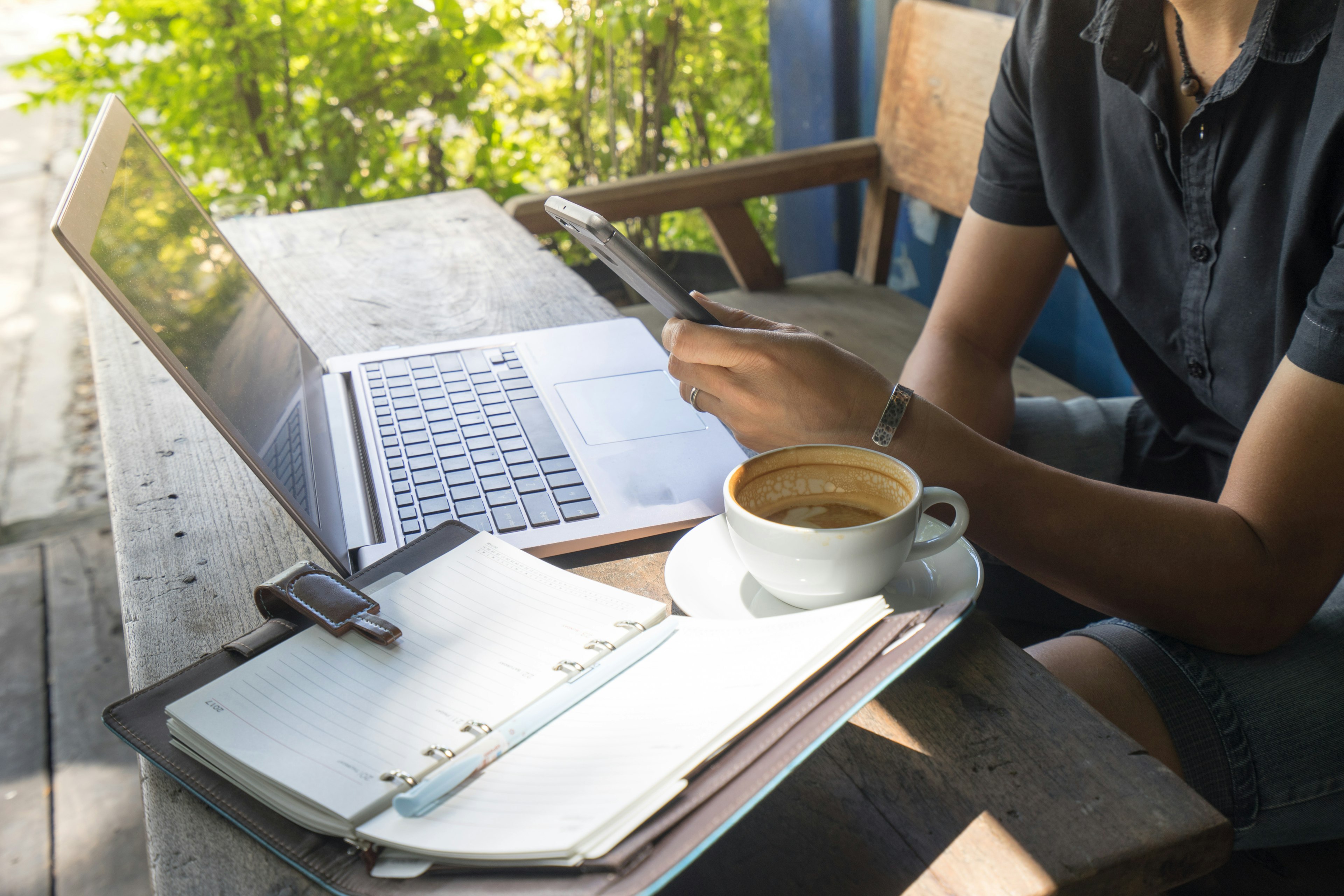 A man is using his phone outdoors at a desk. To his left is a notebook and a half-empty cup of coffee.