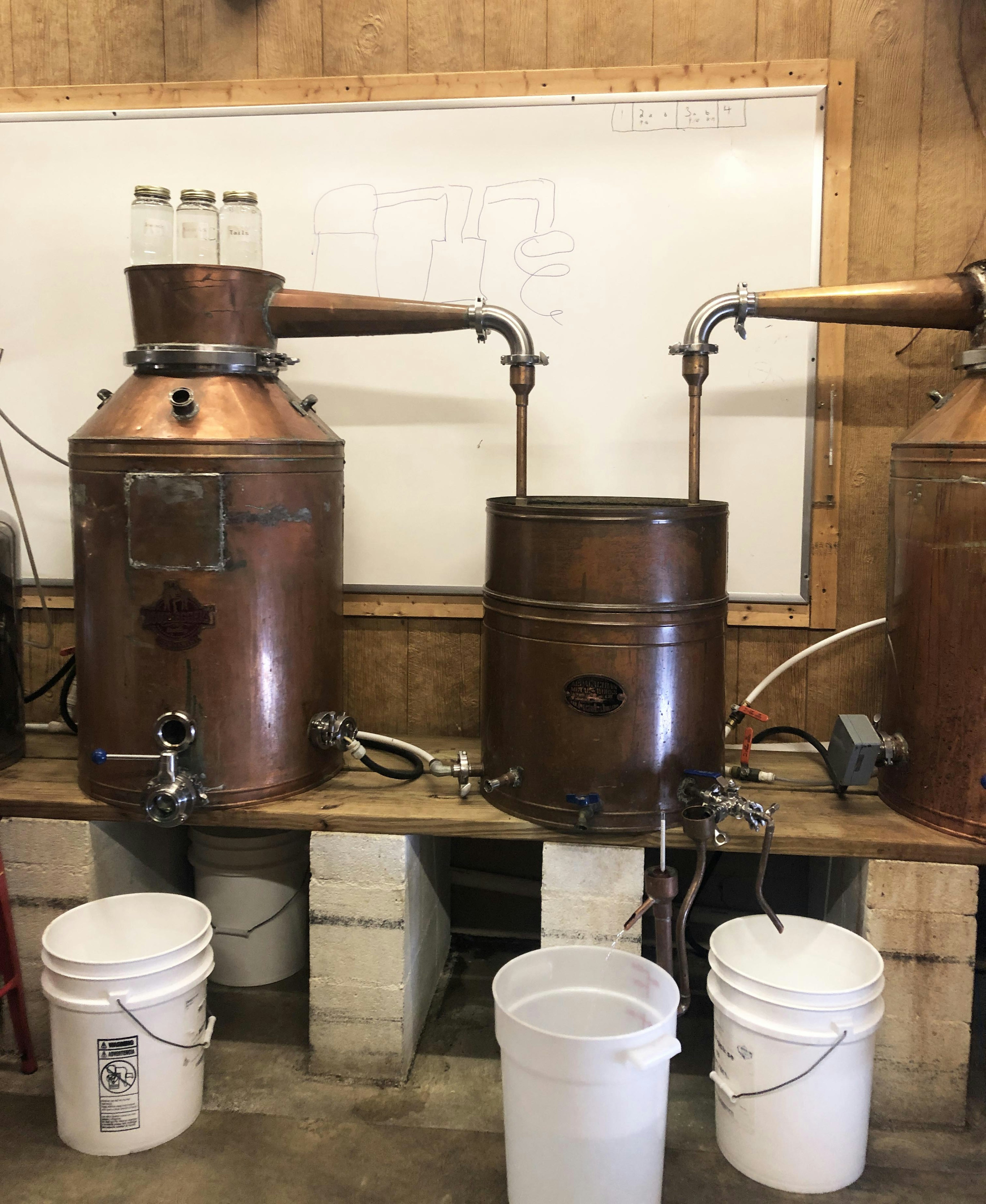 A pair of large copper containers with spouts connected to another large container sit on a raised wooden platform. In front are a trio of white buckets.