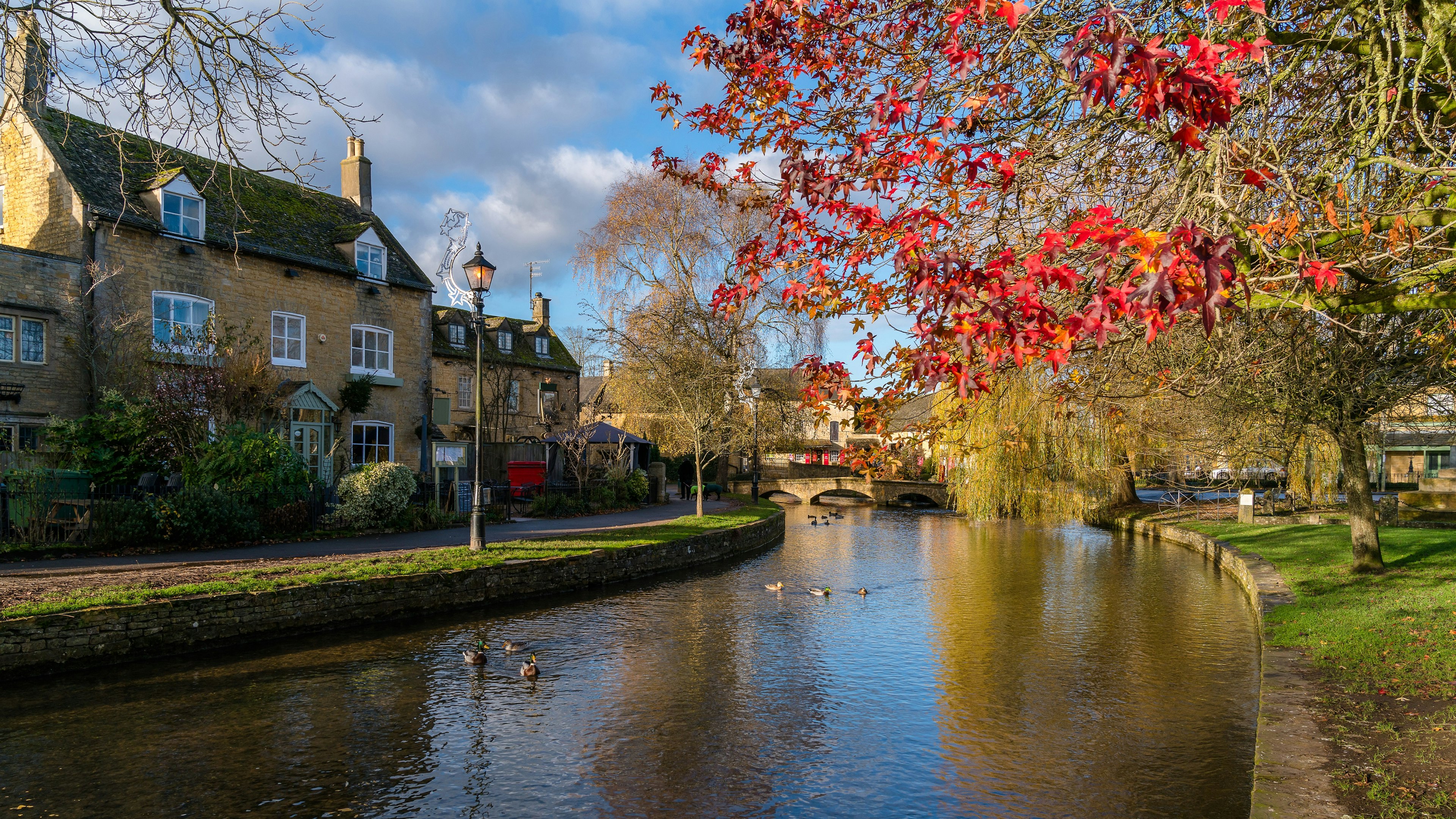 A river meanders past a honey-coloured stone house on one side and a tree bright with red autumnal; leaves on the other.