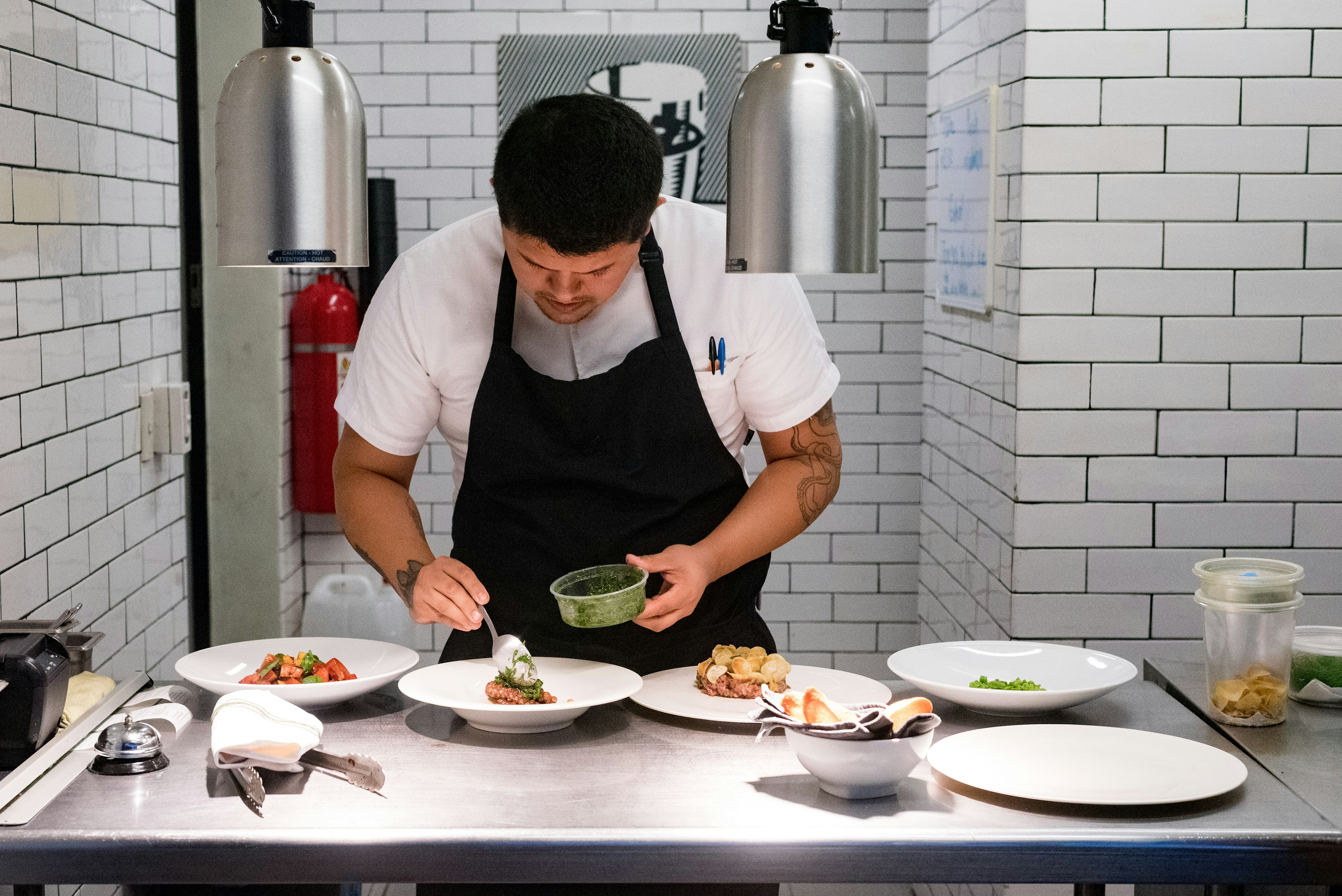 A chef wearing a black apron leans over three white plates, carefully adding a dab of sauce to the dishes