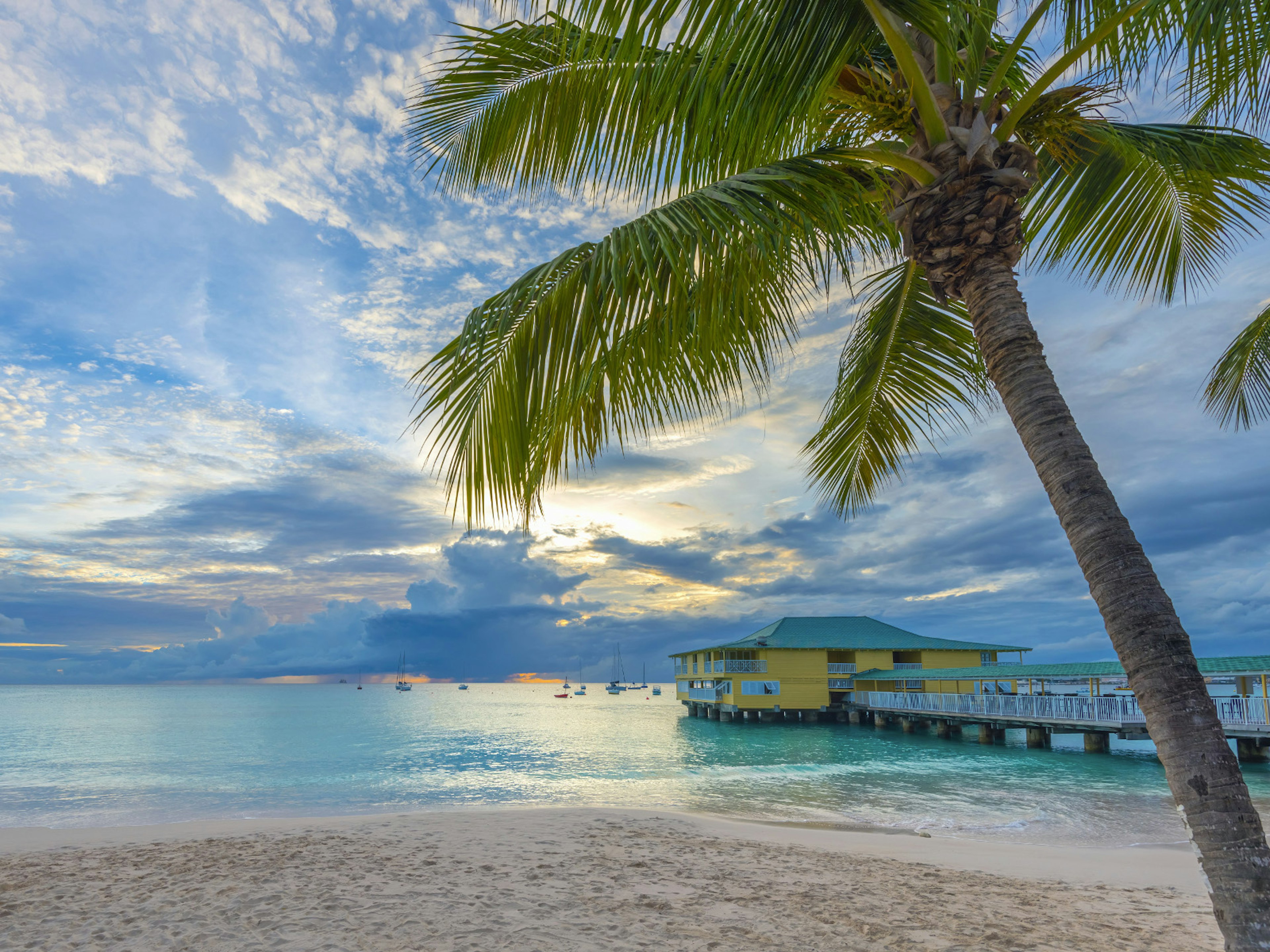 Sunset over Pebbles Beach, Bridgetown, Barbados. A pier stretches over the blue-green sea to a yellow building. There's a palm tree in the foreground leaning over the sandy beach