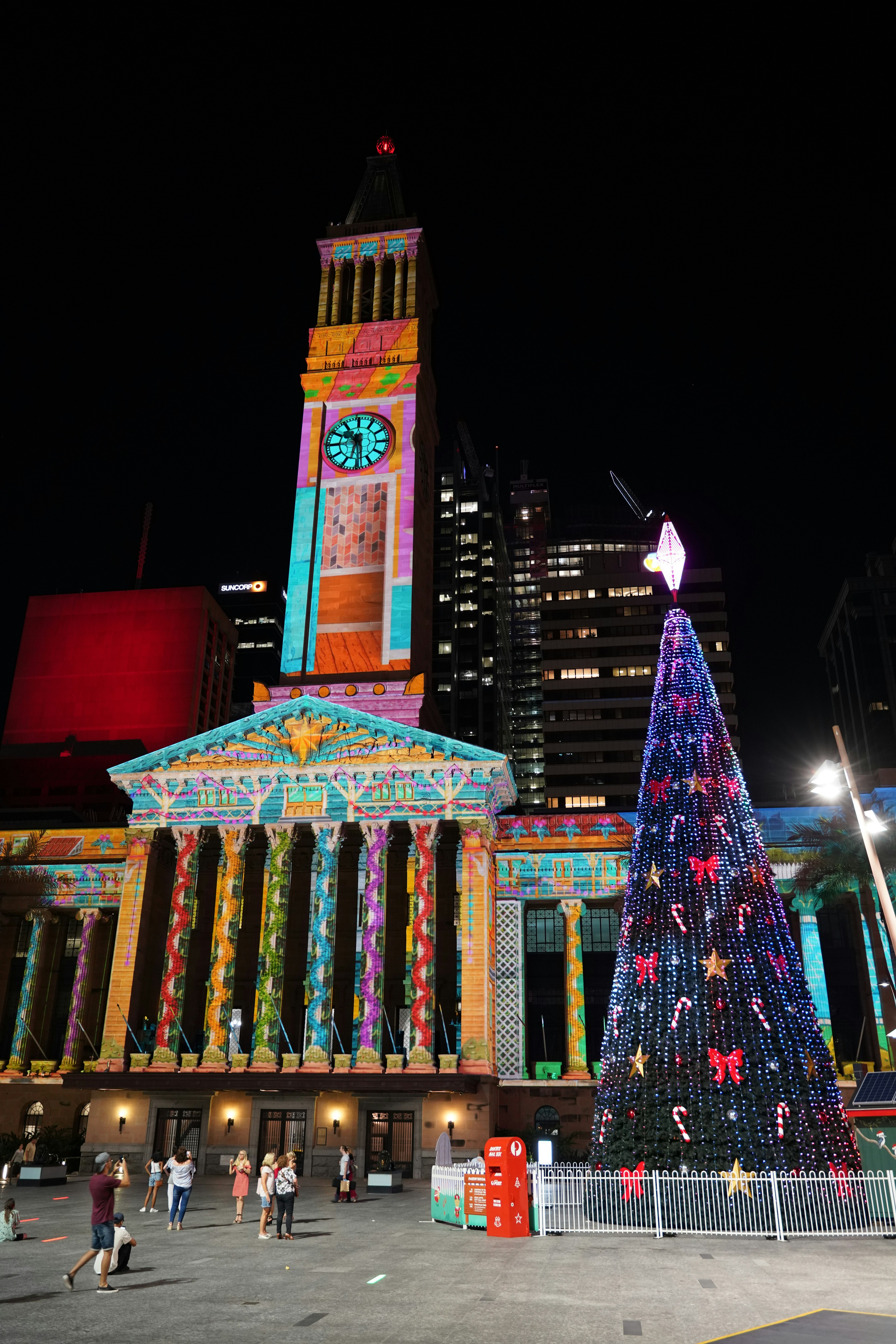 Christmas Lights Show at Brisbane City Hall. The exterior of the hall is illuminated by a colorful light projection and there is a very tall, decorated Christmas tree outside the building