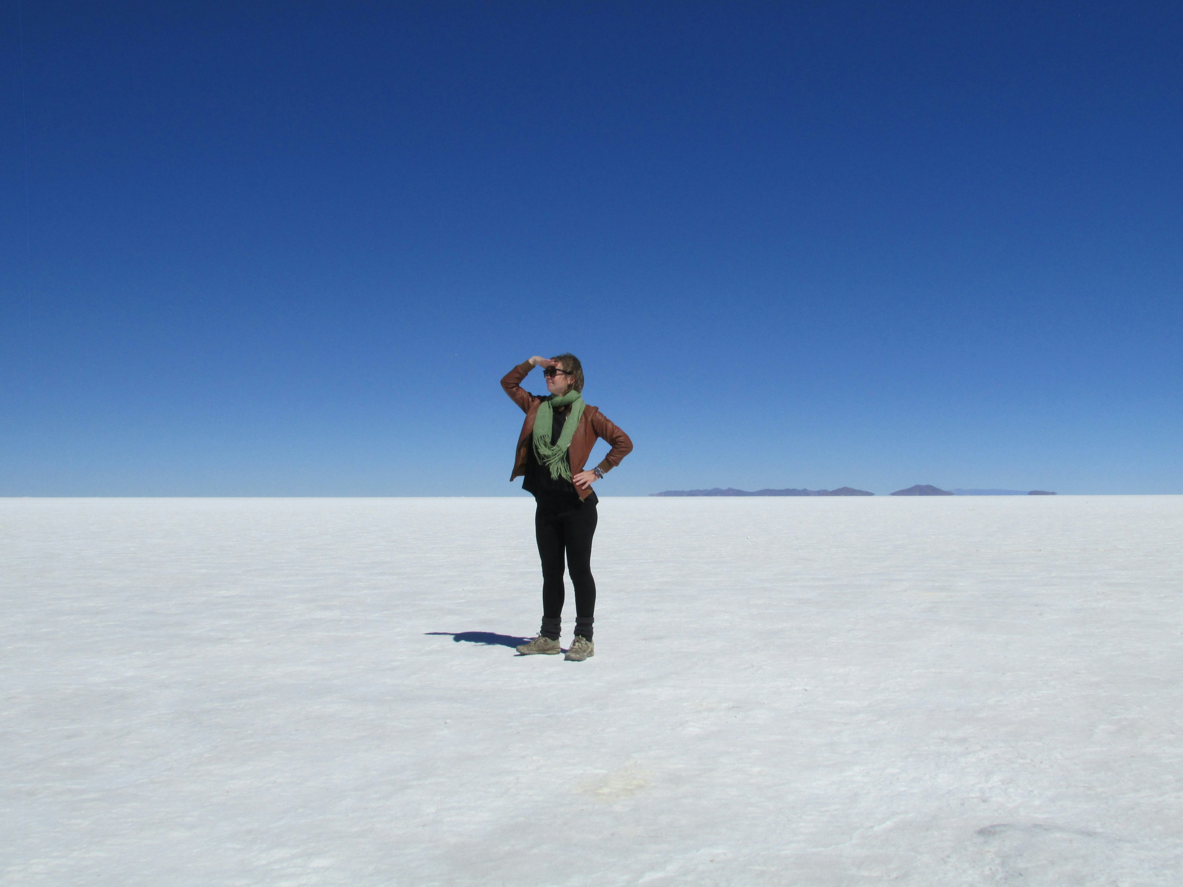 A woman stands in the desert in Bolivia. The sand is bone white and the sky is a pale blue. She is posing by holding her right hand over her eyes.