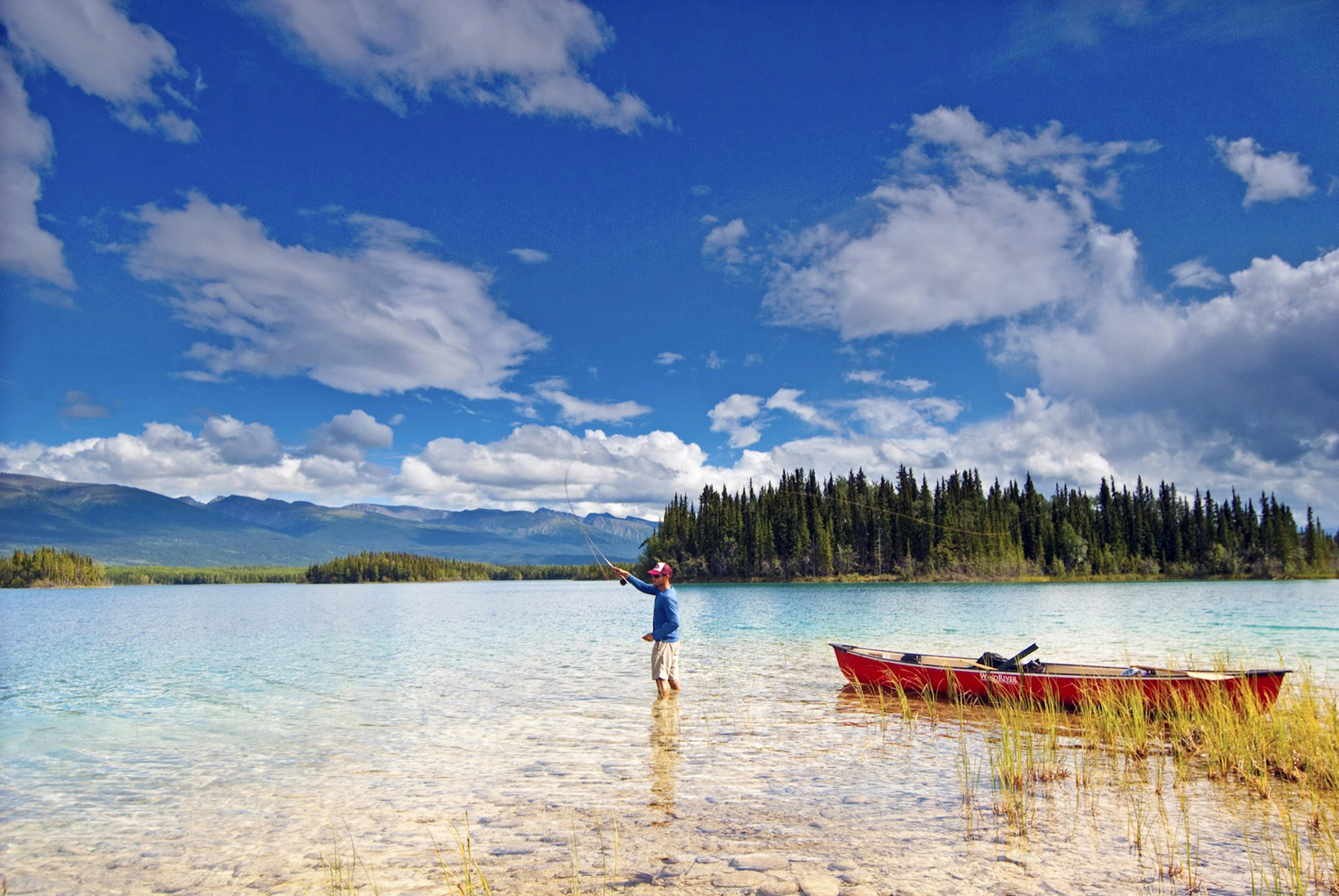 A man in shorts stands in a British Columbia river on a very sunny day, as he casts a fly fishing line next to his red canoe; world's best rivers for fly fishing