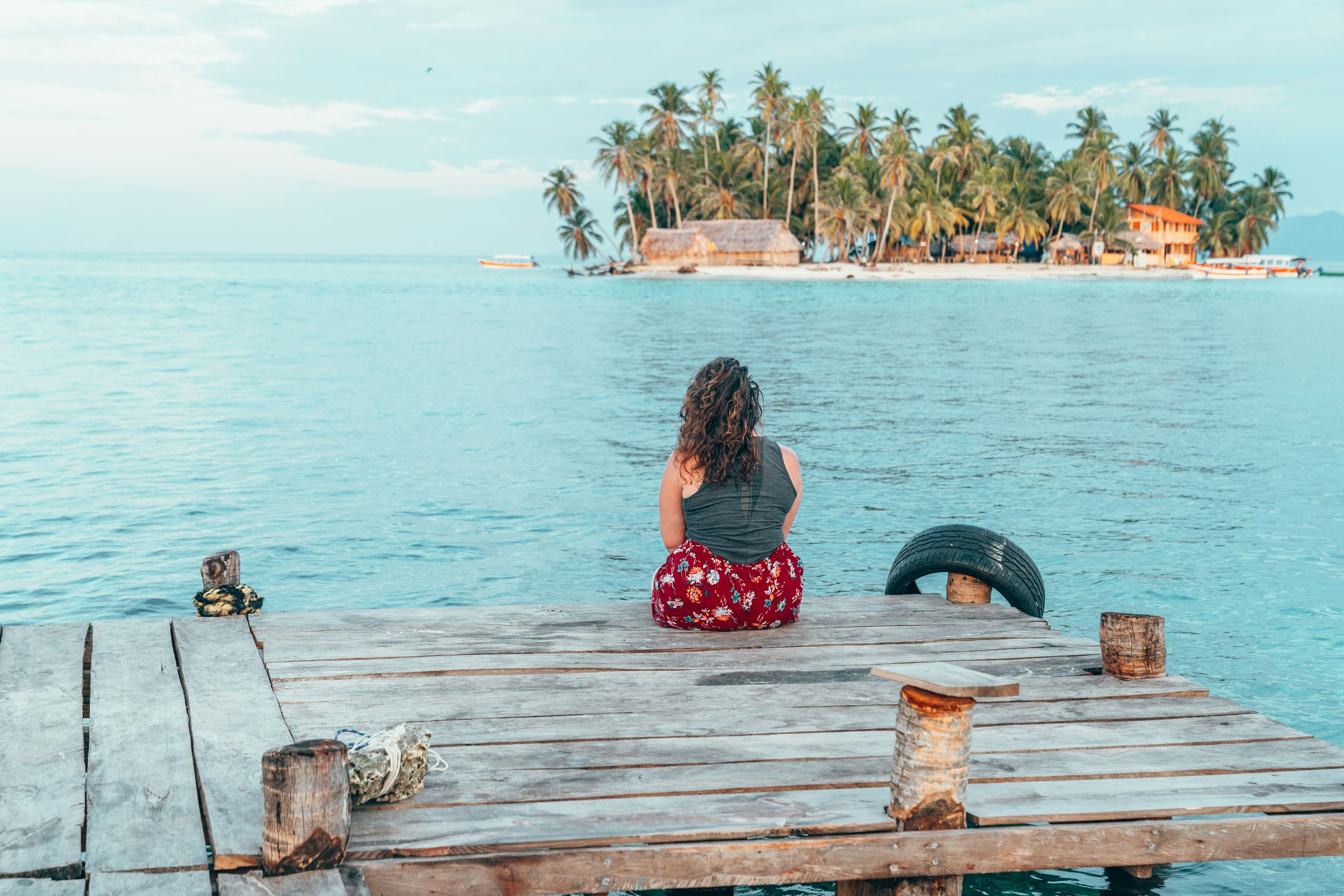 A woman sits on a dock in Thailand, overlooking a blue ocean. An island is within view.