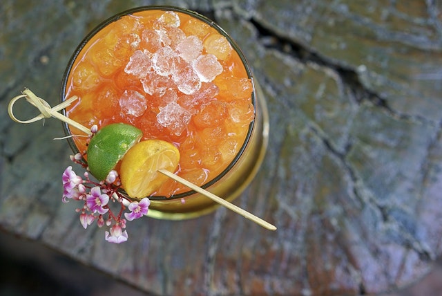 View looking directly down upon a glass containing a bright orange cocktail. The drink is muddled with crushed ice, and a wooden skewer with green and yellow fruits rests artfully across the rim, as well as a delicate sprig of flowers.