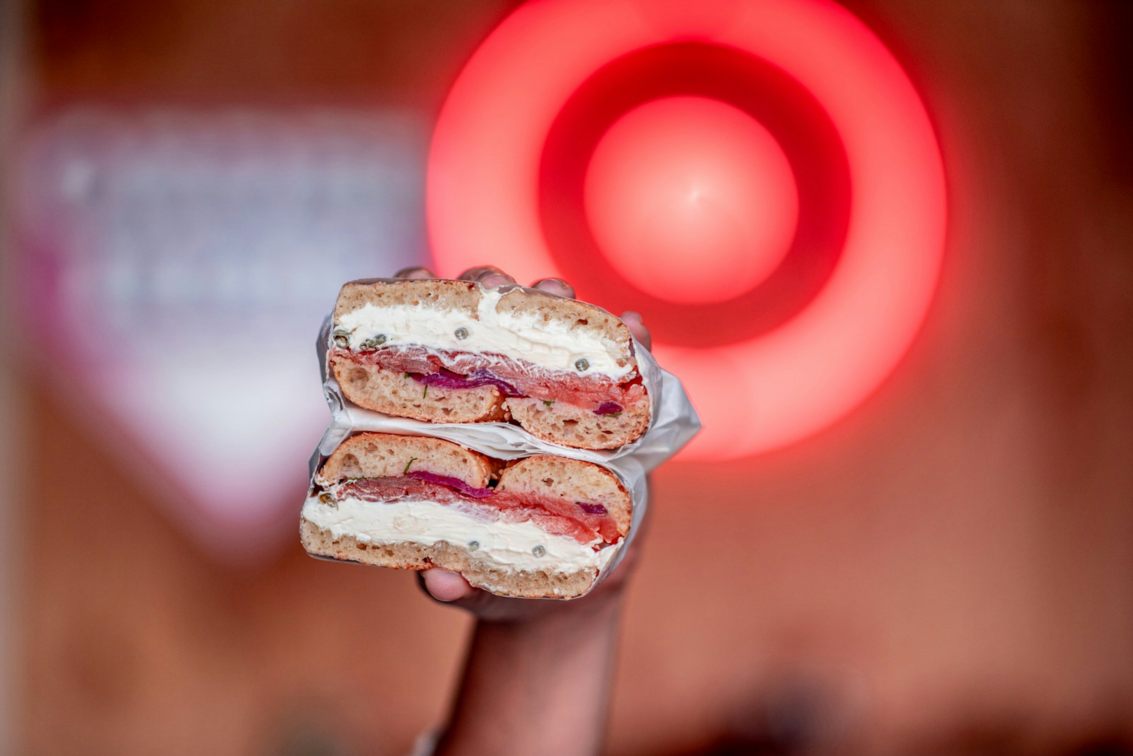 A close up of a hand holding a bagel, halved and wrapped in white grease-proof paper. The bagel is filled with cream cheese, capers, tomato and red onion.