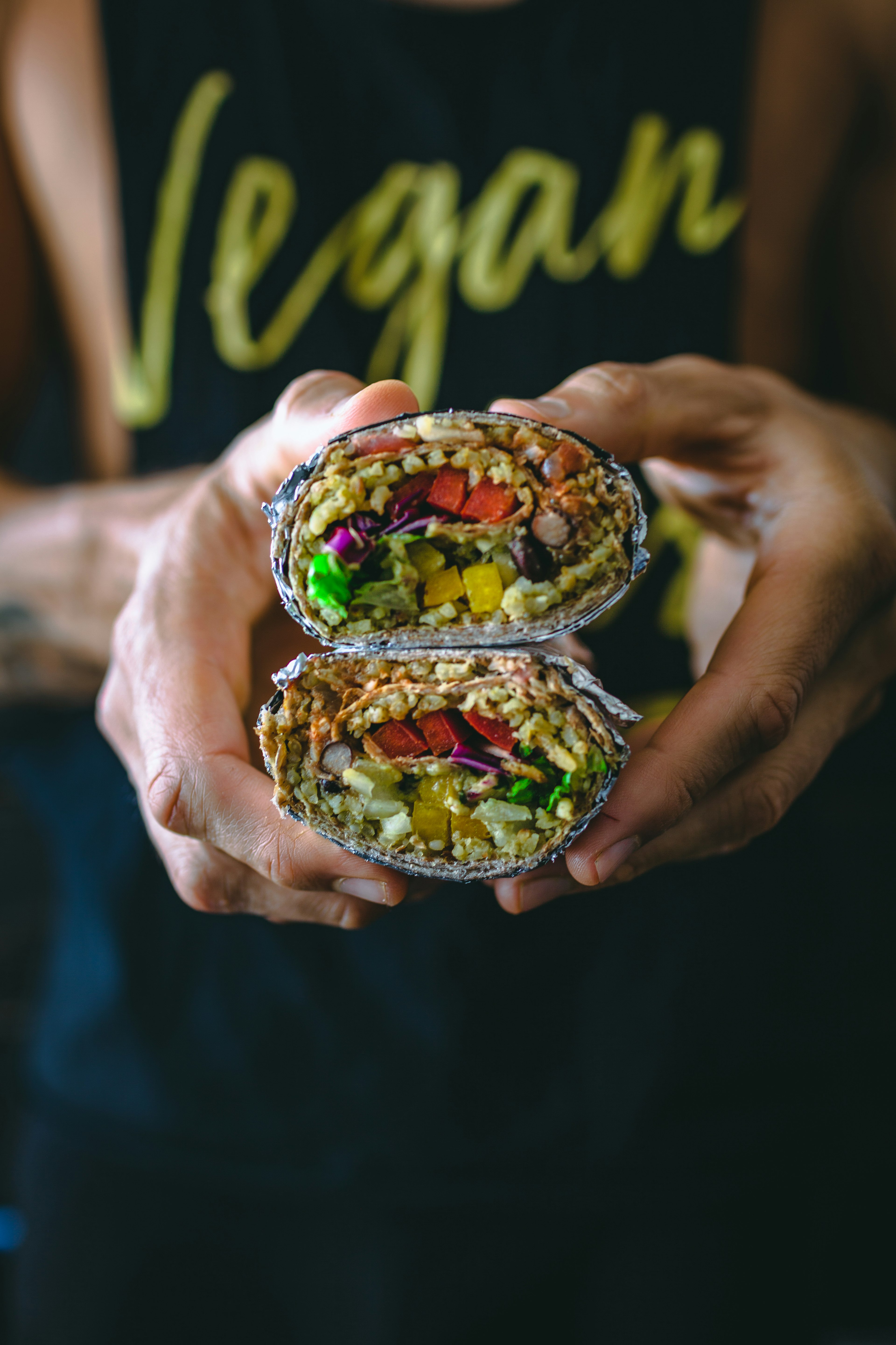 A man wearing a t-shirt that says 'vegan' holds a burrito filled with vegetables towards the camera