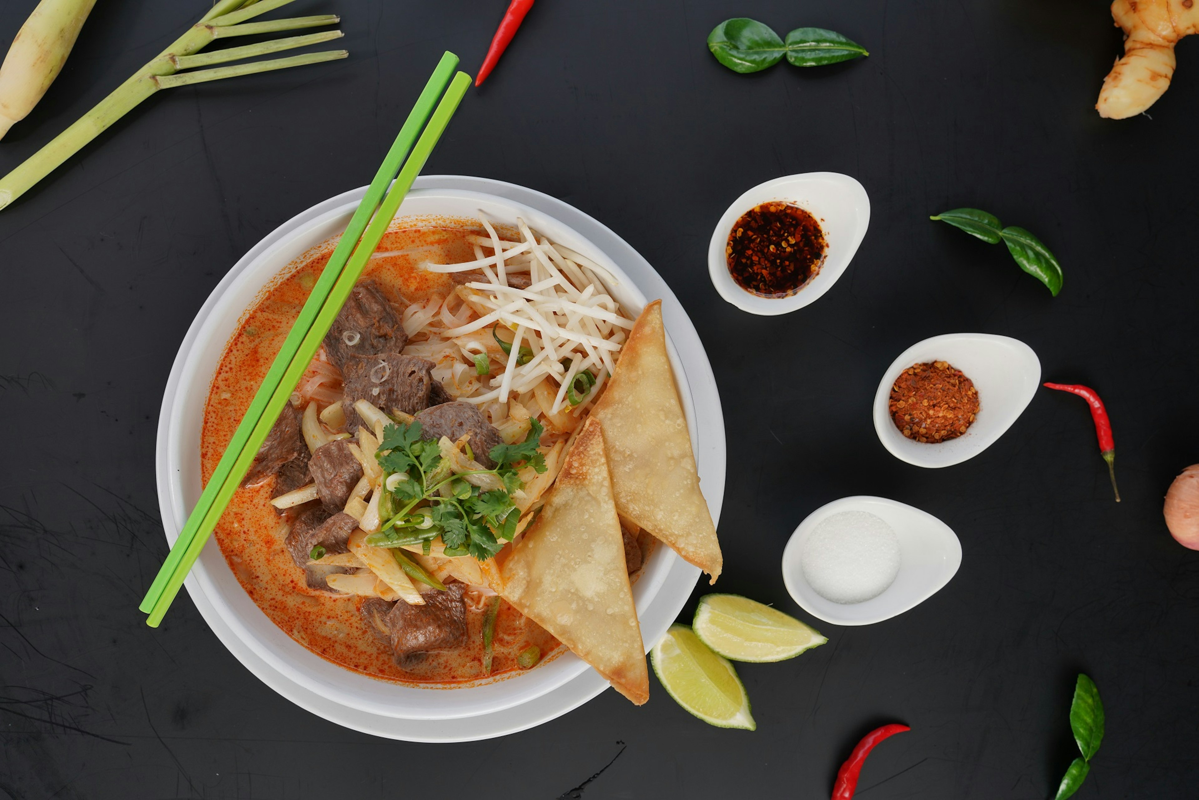 Overhead view of a spicy vegan noodle dish. There are a pair of chopsticks positioned across the side the bowl. There are spices and sauces placed around the bowl; LA vegan restaurants