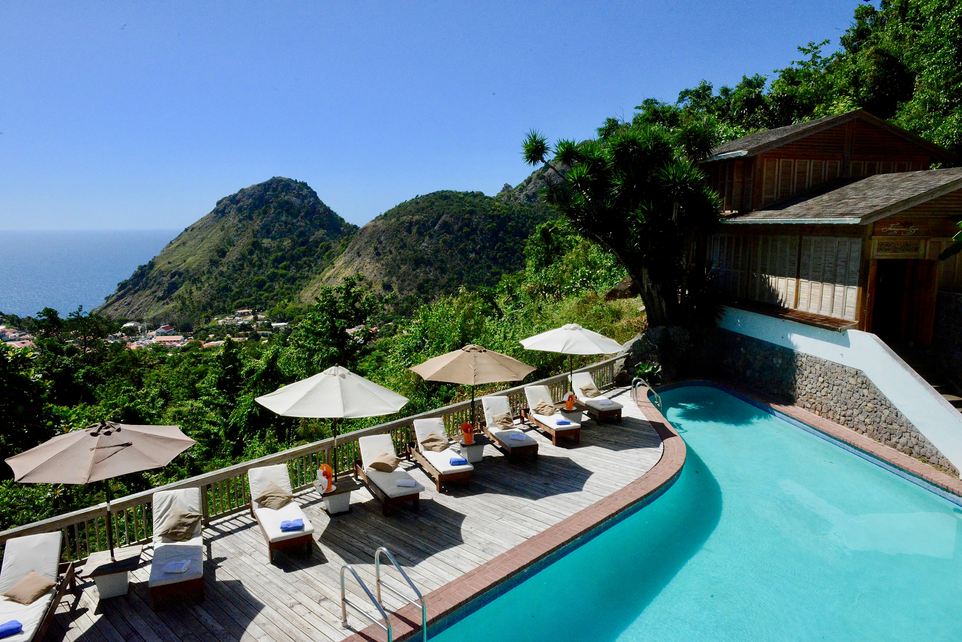 Deck chairs are lined alongside a pool at Queen's Gardens Resort and Spa. Behind a fence a lush, green valley is visible, next to two mountains.