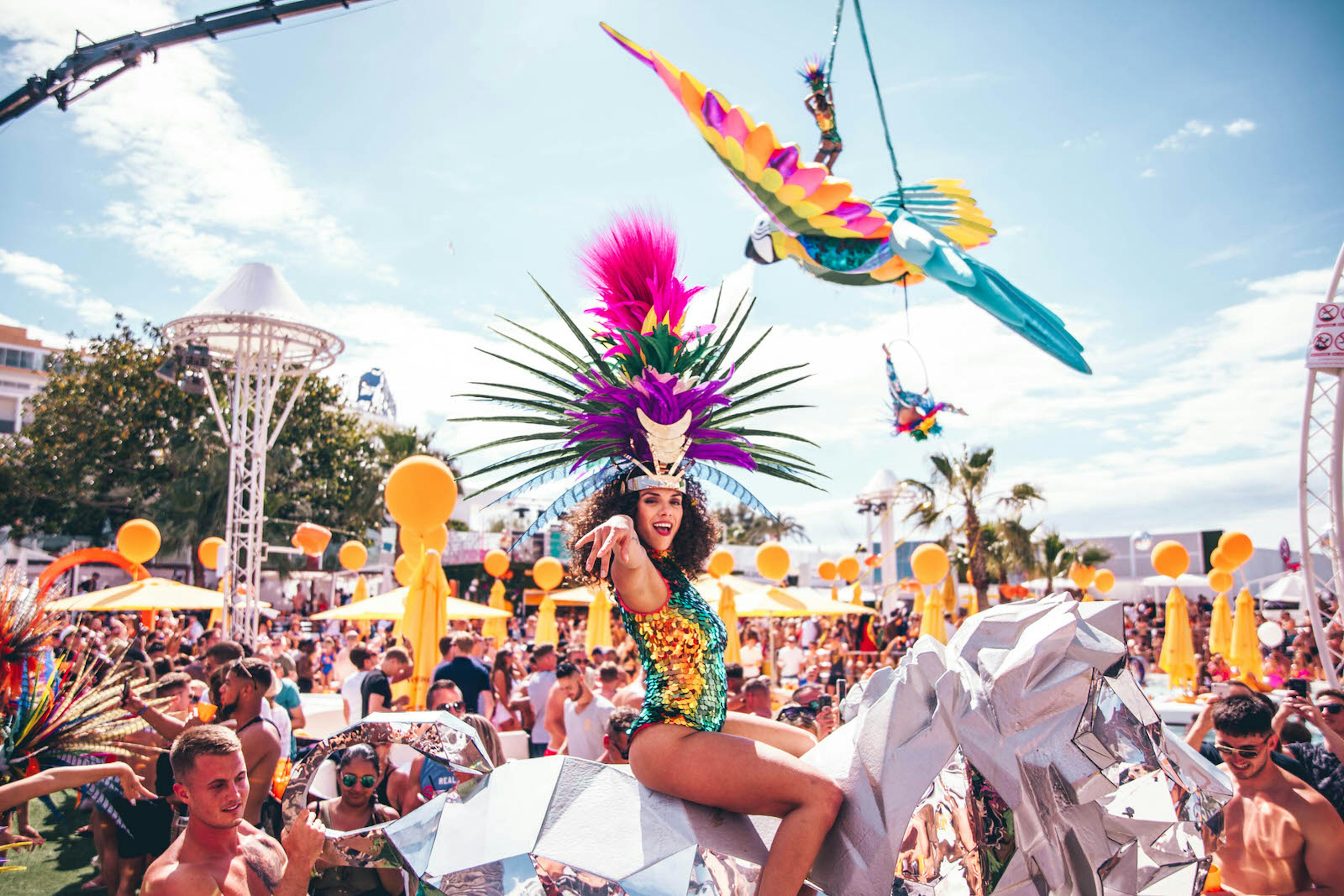 A woman in a colourful sequined leotard and a huge multicoloured feather headdress sits on top of a silver unicorn in front of a huge crowd at a pool party in O Beach Ibiza during the day