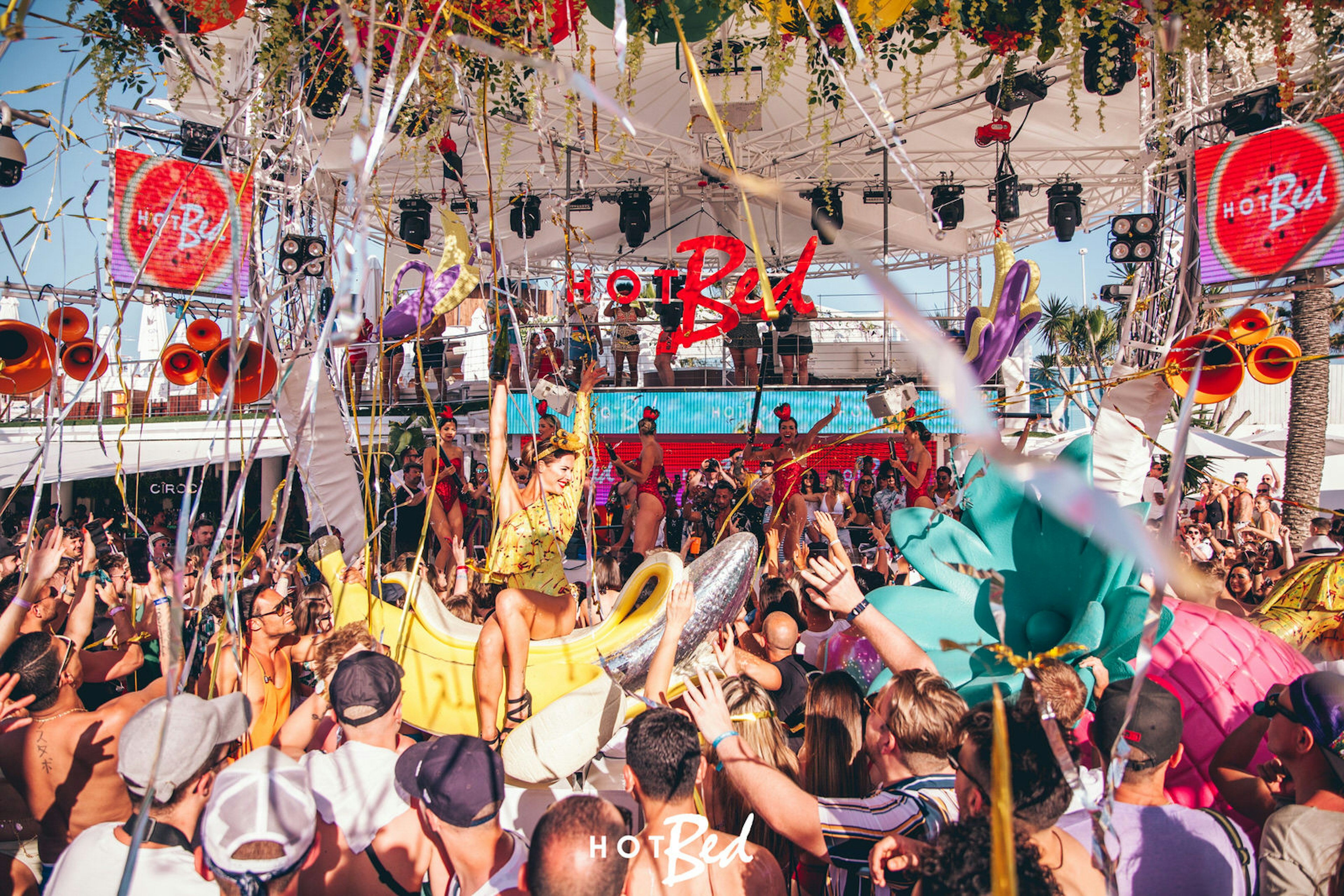 A crowd are gathered around a giant plastic banana with a performer sitting on it with their hands in the air. There are silver streamers in the air and a covered stage in the background.