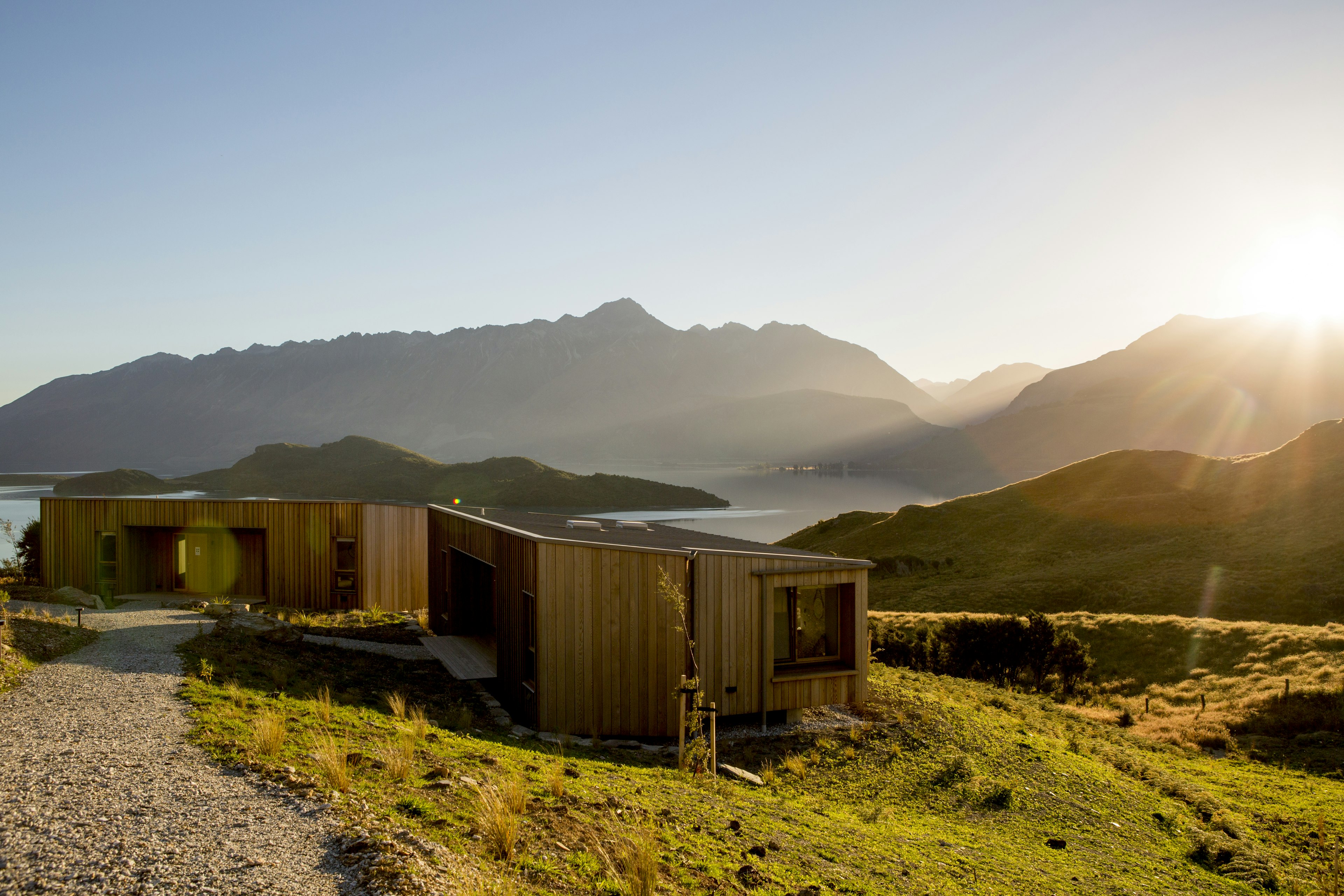 Simple but stylish wood cabins on a grassy plateau amidst rugged scenery beside a lake. The white-hot sun is low in the sky and the photo has captured its rays.