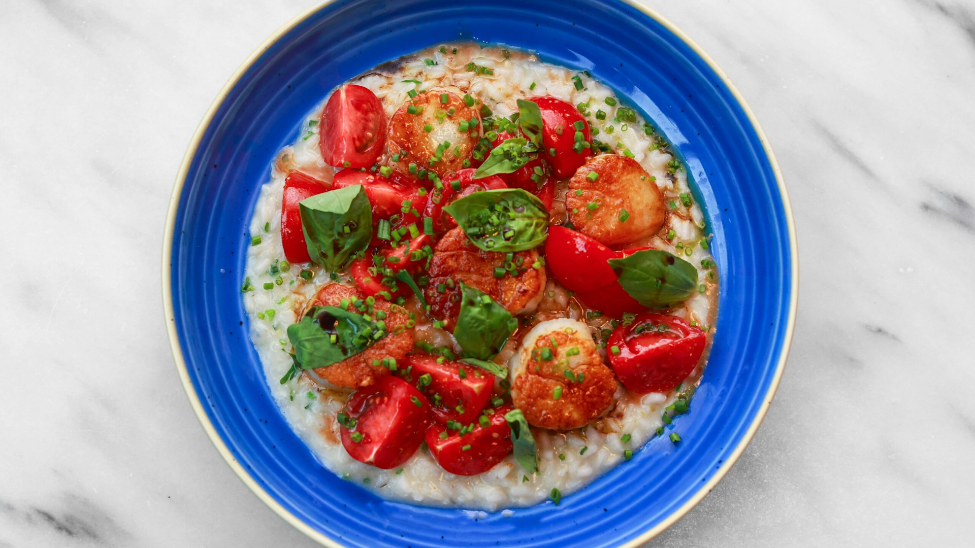 An overhead view of seared scallops, red tomatoes and basil leaves on a bed of risotto in a blue bowl. 