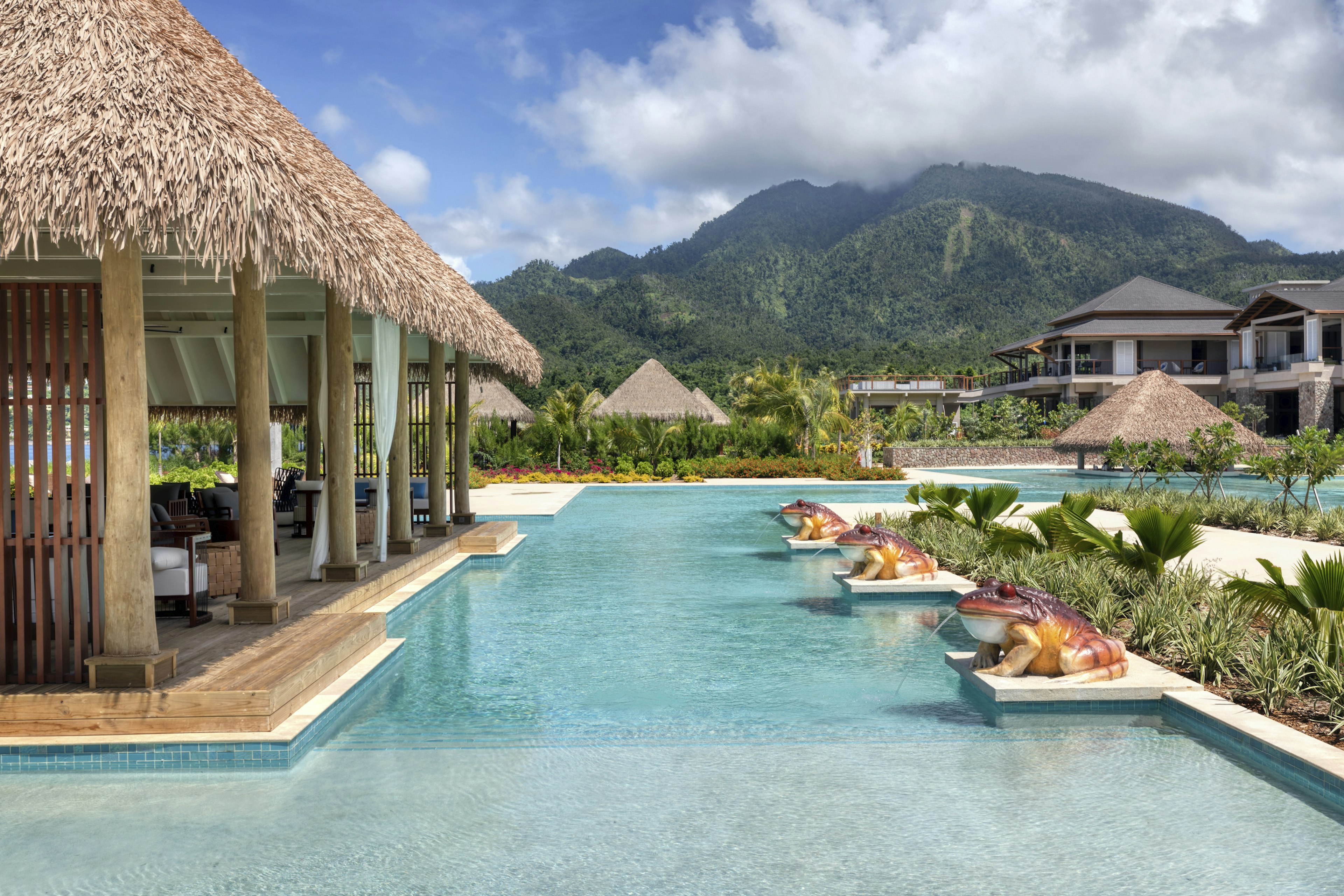 Exterior shot of Cabrits Resort's pool with three large frog statues spitting water into the large pool. There is a large open-air thatched-roof common area filled with chairs and tables. A patch of clouds cover the peak of a large verdant mountain in the distance.