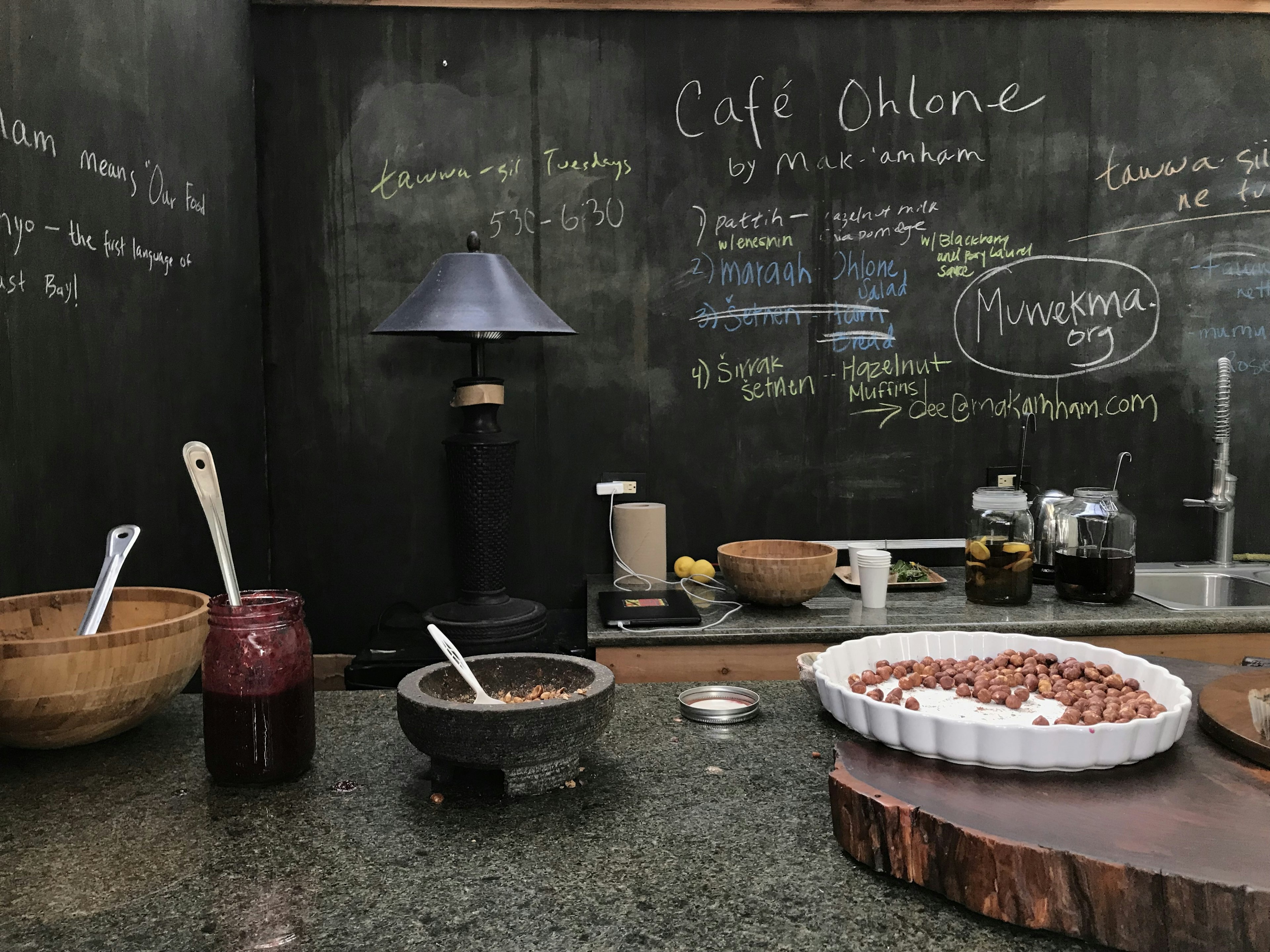 A blackboard is covered with several messages. On the front counter, there are wooden bowls, a jar of jam and mortar filled with nuts; Indigenous food