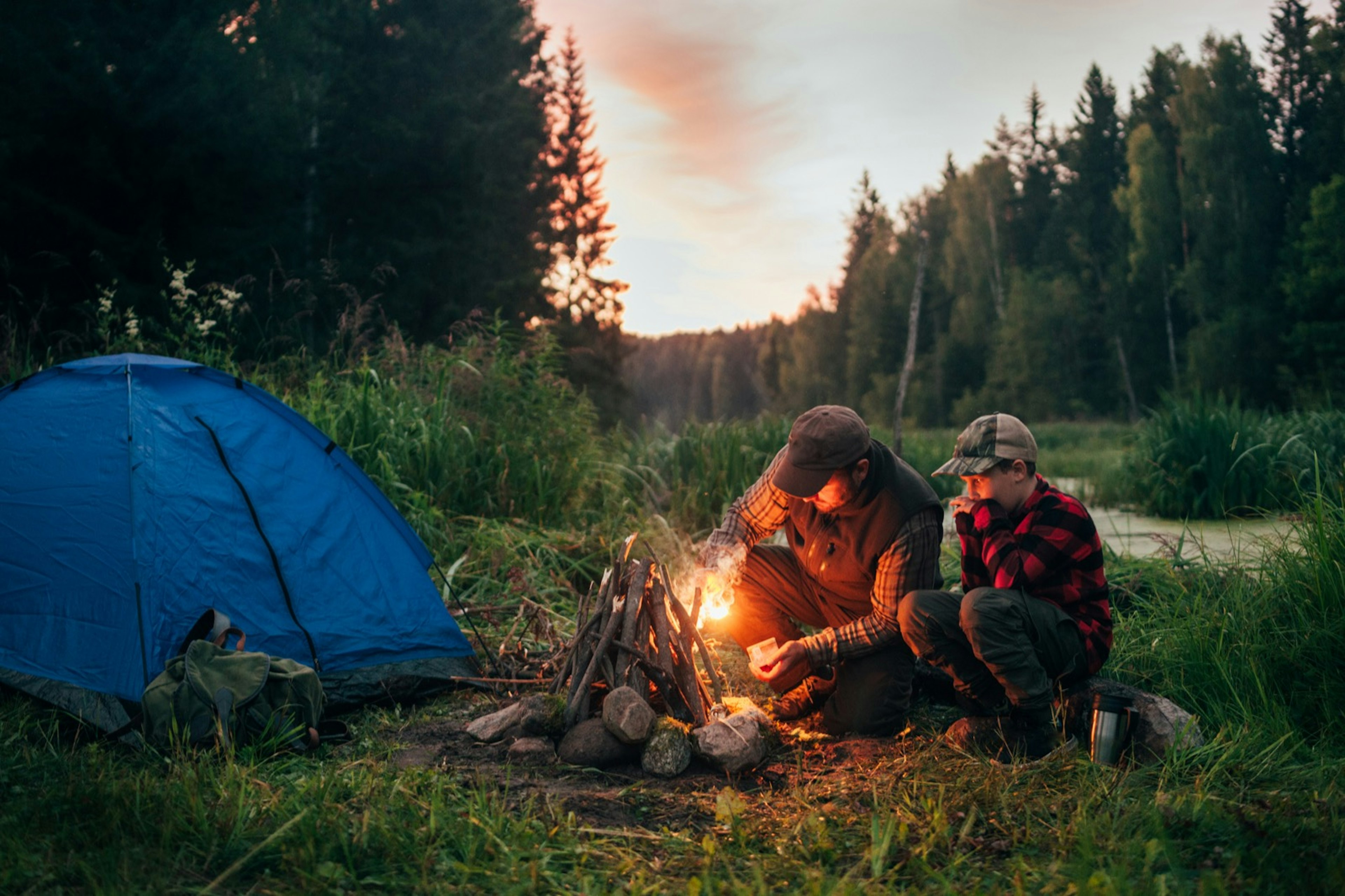 A man and son start a fire as the sun begins to set in the woods of coniferous trees. A small blue tent is near the campsite. They both wear peaked hates and checked shirts.