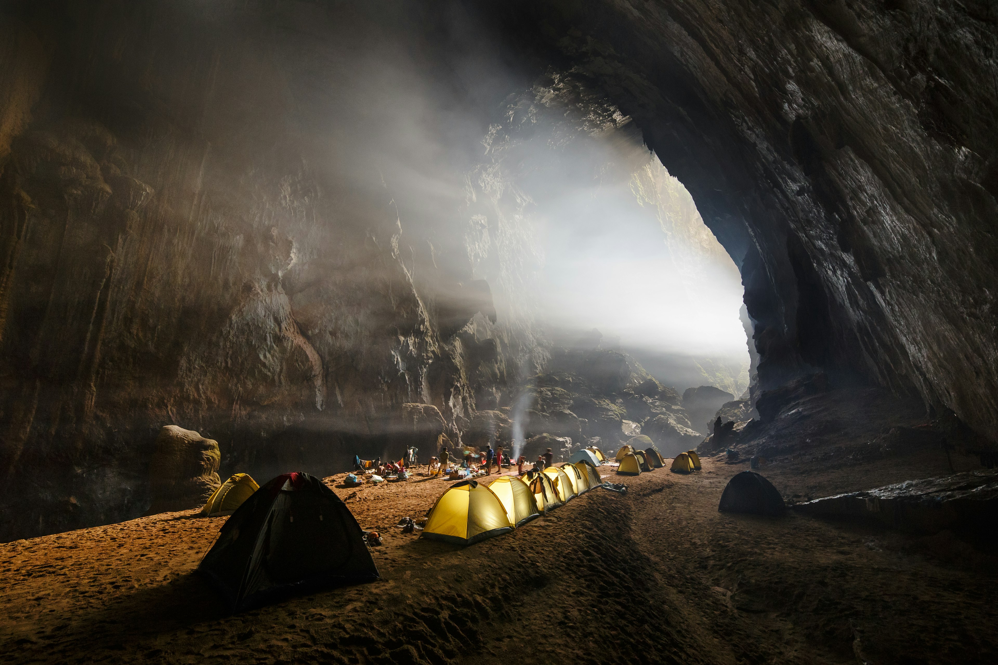 A row of tents is lined up on the rock inside Hang Son Doong cave, while sunlight beams in through an opening.