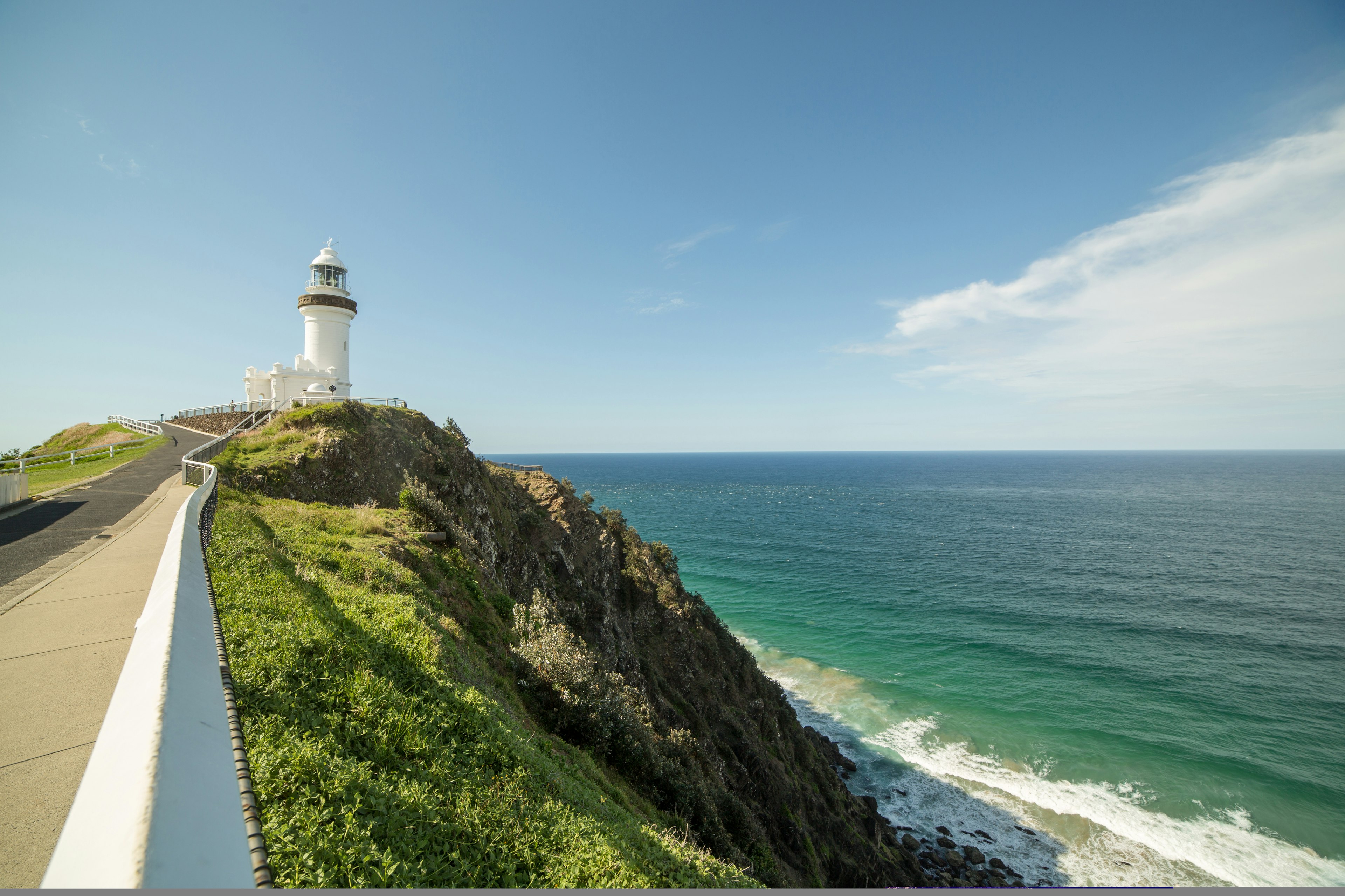 A road leads towards a white lighthouse at the end of the Cape Byron Walking Track; it stands on a rocky cliff, with waves crashing beneath.