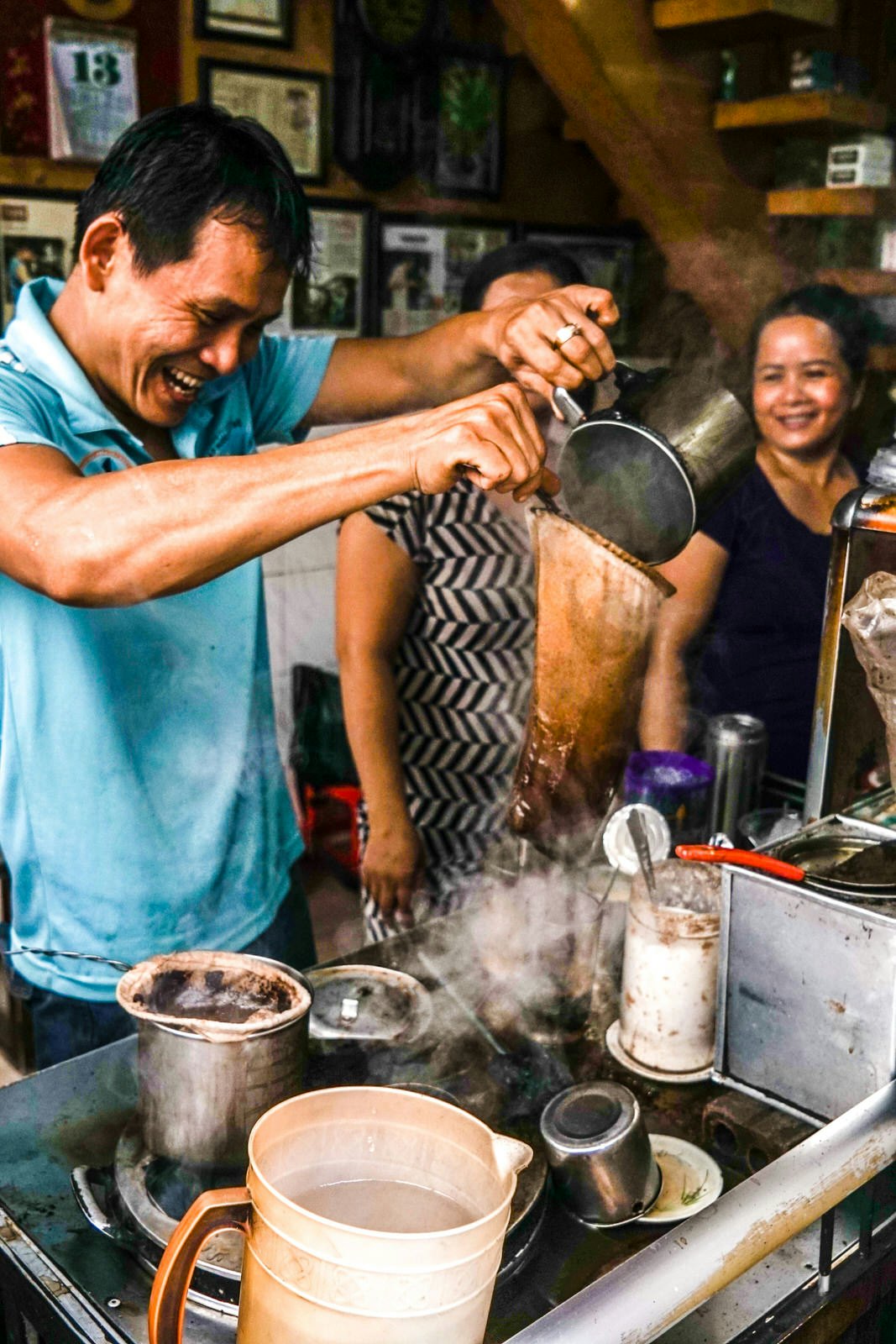 Man filters coffee through a net-like material in a Vietnamese cafe, everyone's smiling and laughing from the caffeine highs